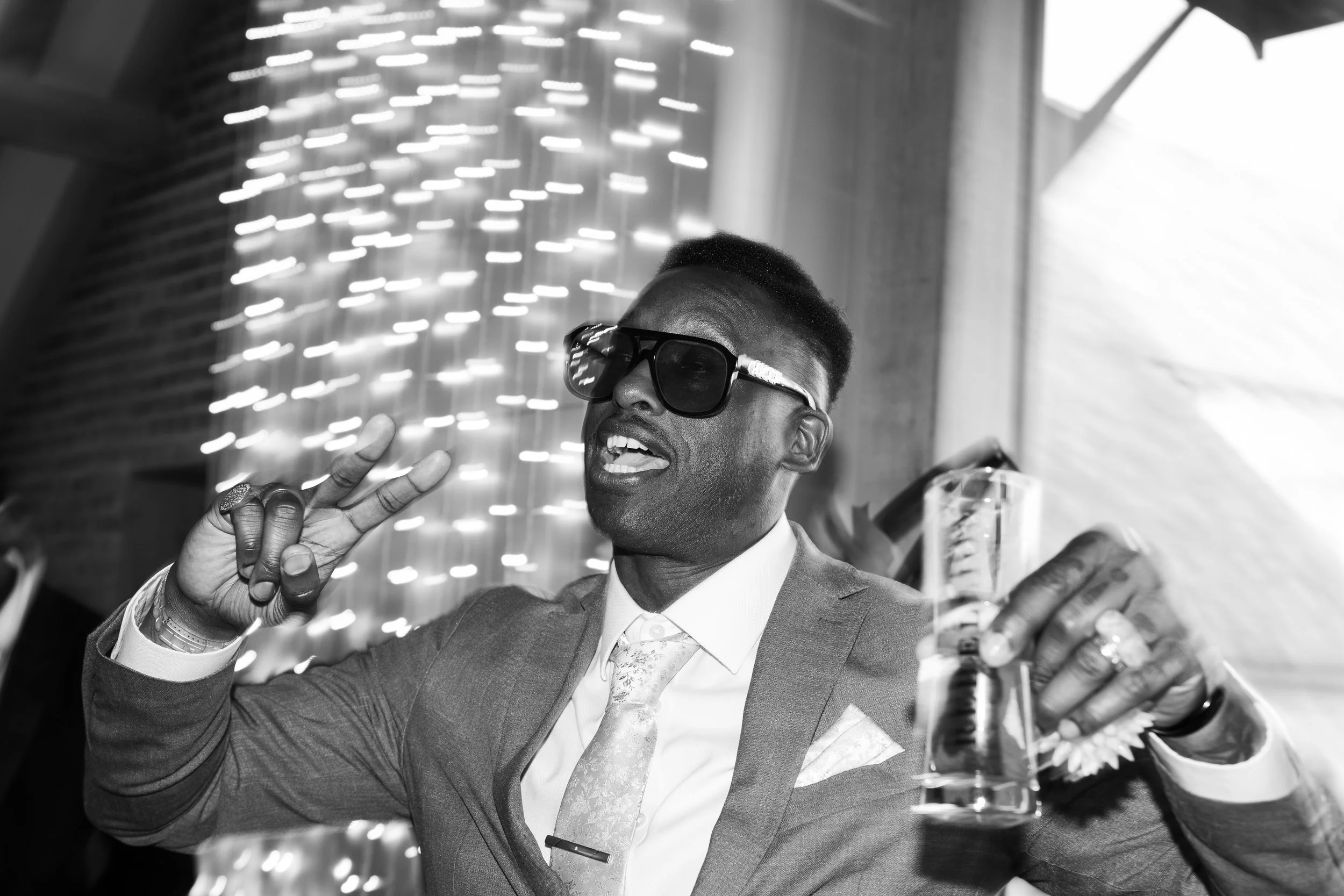 A man wearing a suit and sunglasses holding a drink, making a peace sign with his fingers at a party or celebration.