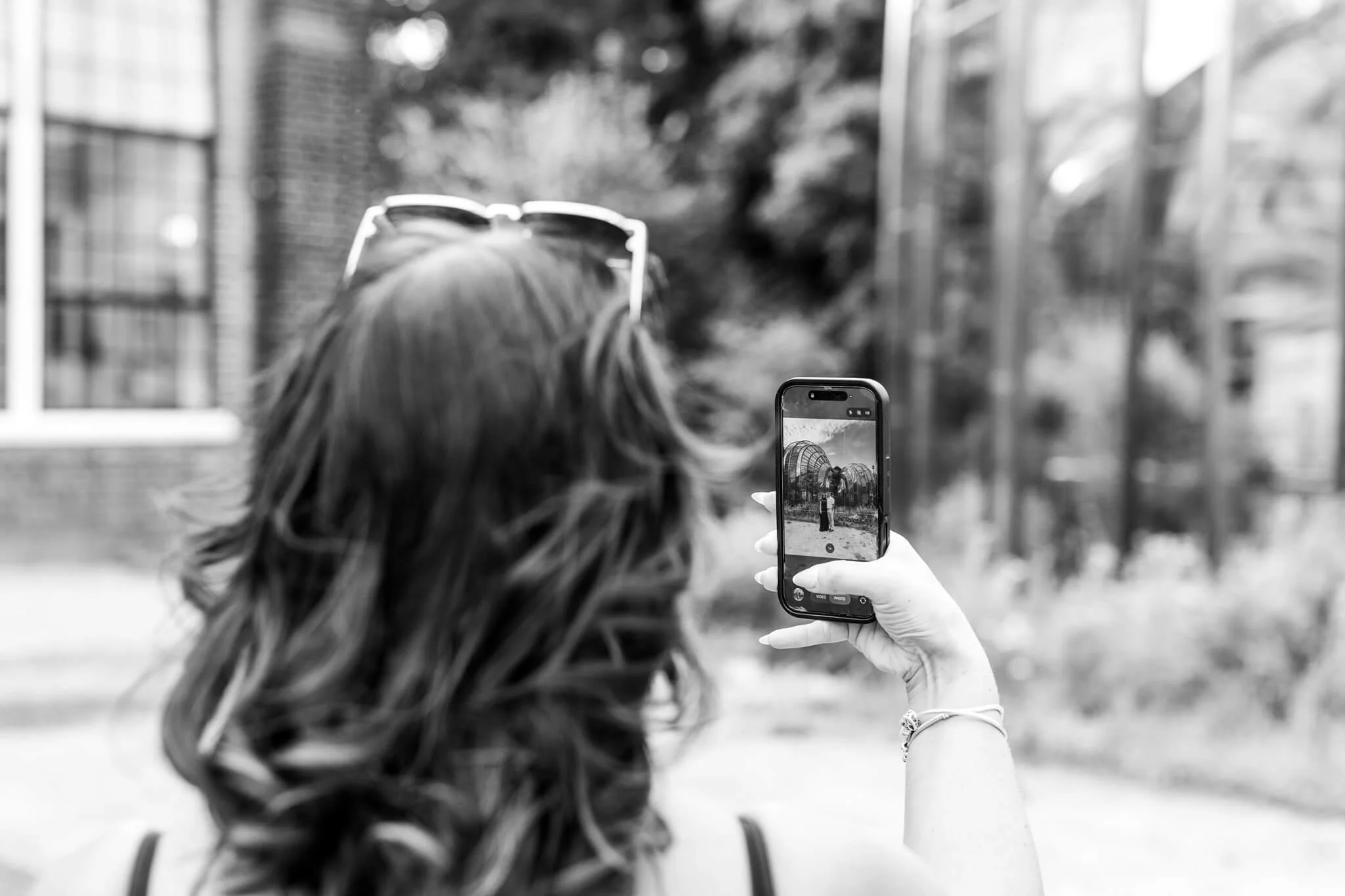A person with long hair and a bracelet is taking a photo with a smartphone of another person standing outdoors near a structure, with trees and buildings in the background. The photo is in black and white.