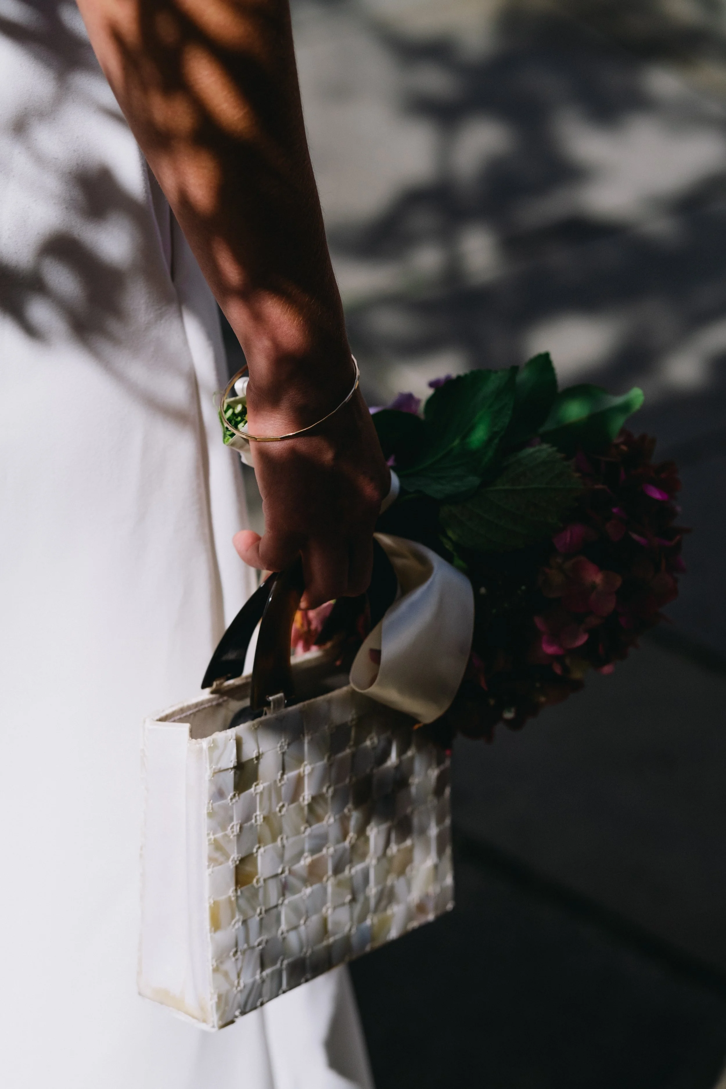 Close-up of a person holding a white handbag with a lattice design, a bouquet of flowers inside, and wearing a gold bracelet, with sunlight and shadow patterns on their arm.