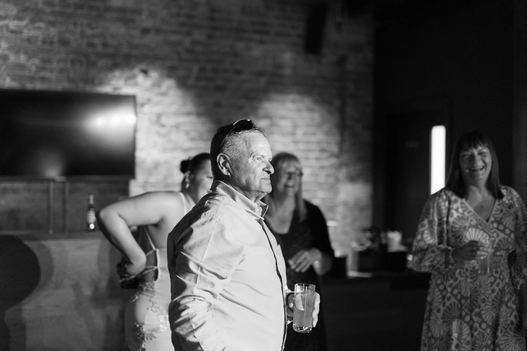 A group of people standing in a room with brick walls, enjoying a social gathering or celebration. One man in the foreground is holding a drink and smiling, while others are seen smiling and engaging in conversation.