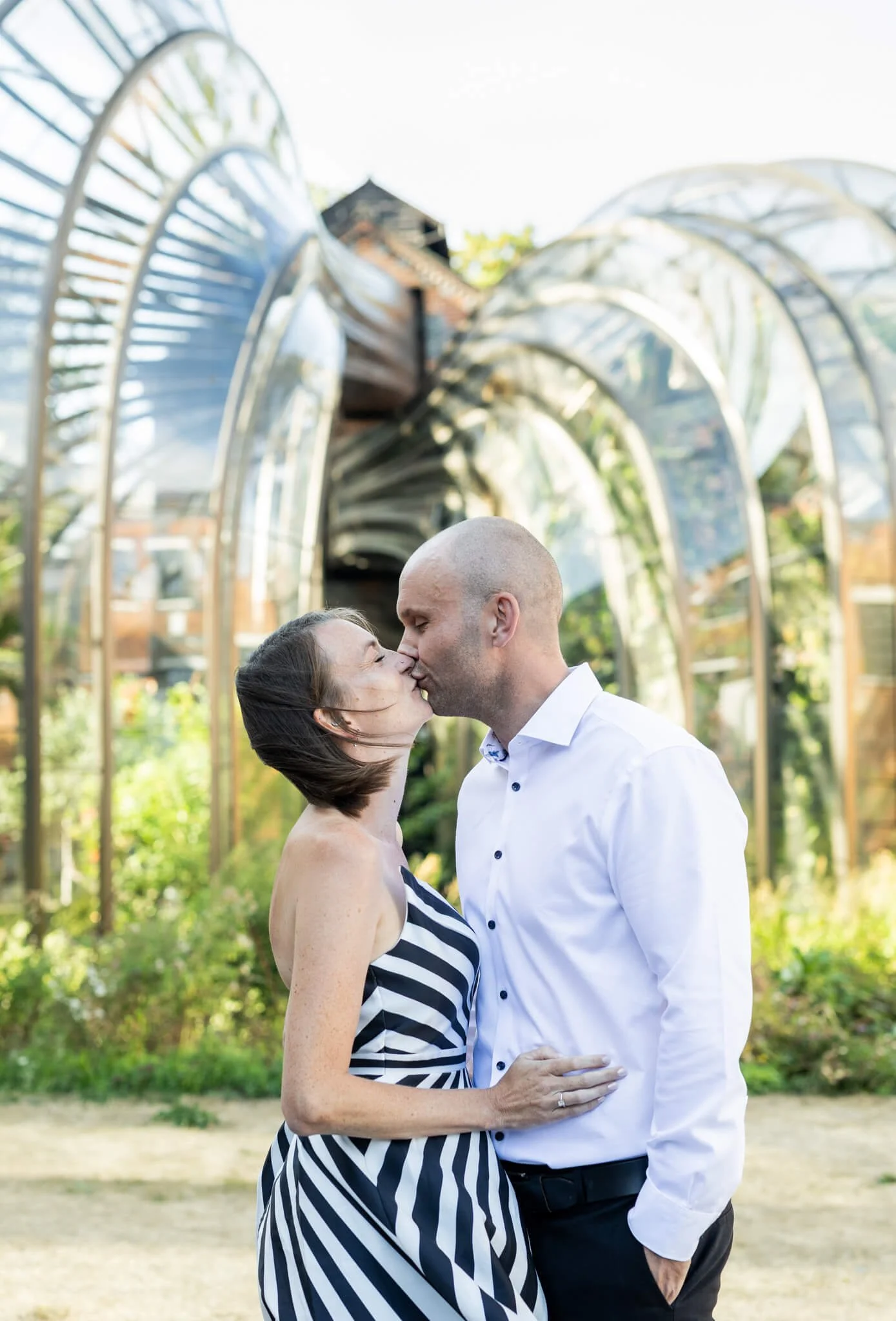 A couple kissing outdoors in front of a reflective, arch-shaped metal sculpture, with trees and greenery in the background.