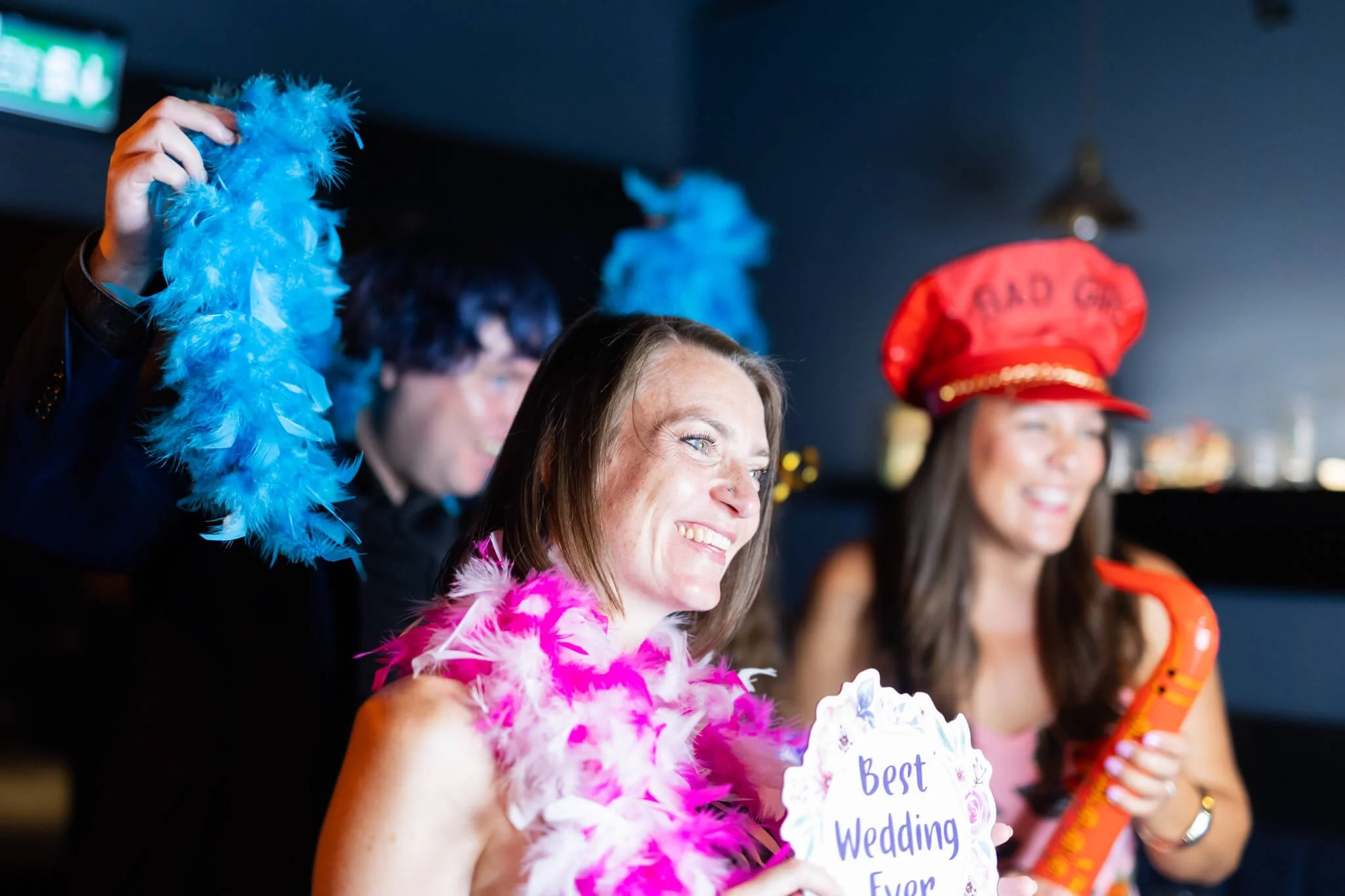 A group of women at a celebration, wearing colorful feather boas and holding signs. One woman in the foreground is smiling and holding a sign that says 'Best Wedding Ever,' wearing a pink feather boa. In the background, two women are visible, one wit