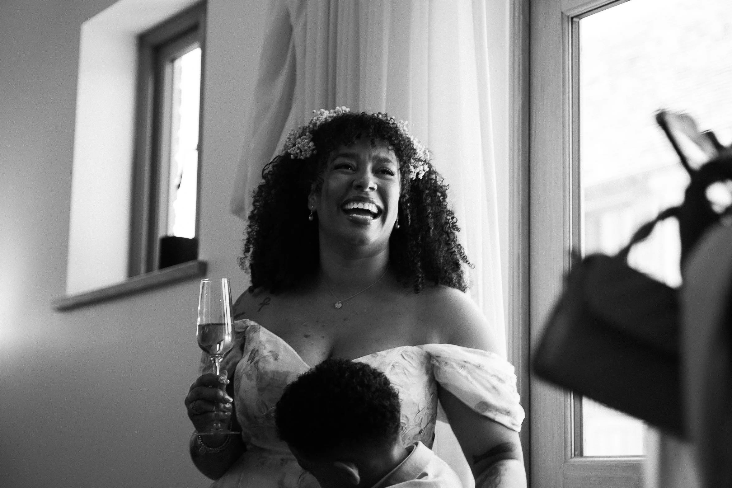 A woman with curly hair and a flower crown smiling in front of a mirror, holding a glass of champagne. She is wearing a floral dress and earrings, and appears to be at a celebration or special event.