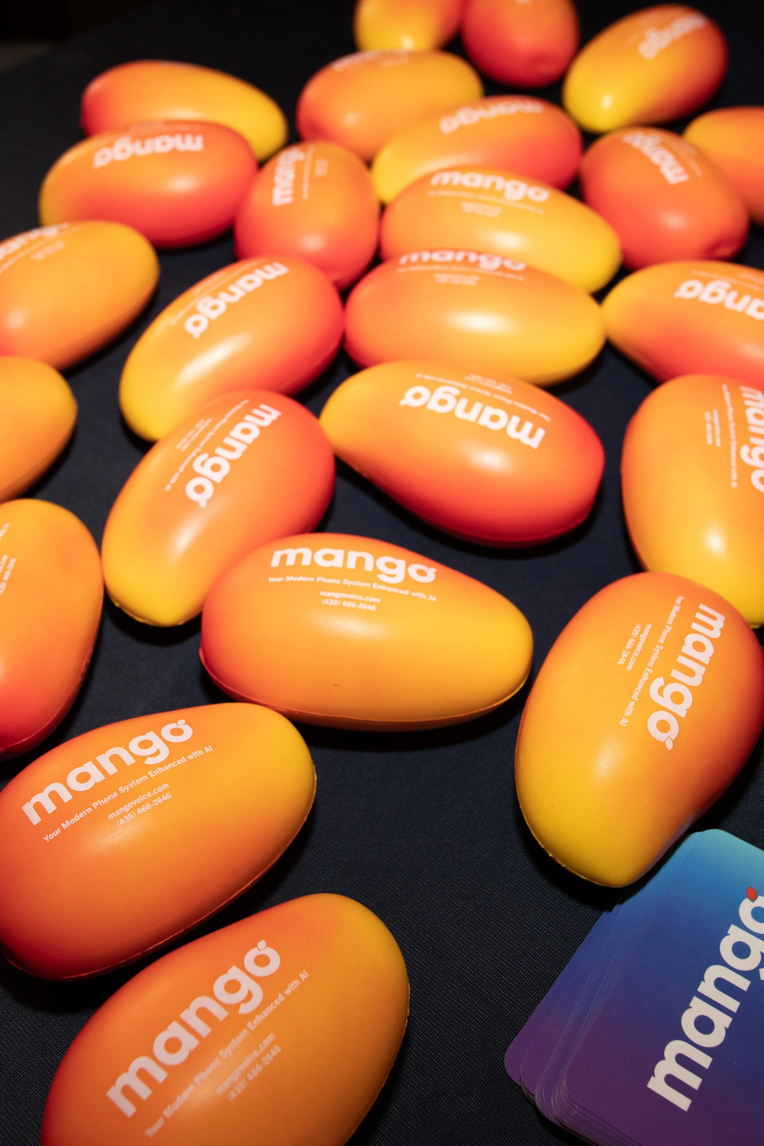 Multiple orange and yellow mango-shaped objects with the word 'mango' printed in white on their surfaces, arranged on a black surface alongside a stack of purple cards with the same branding.