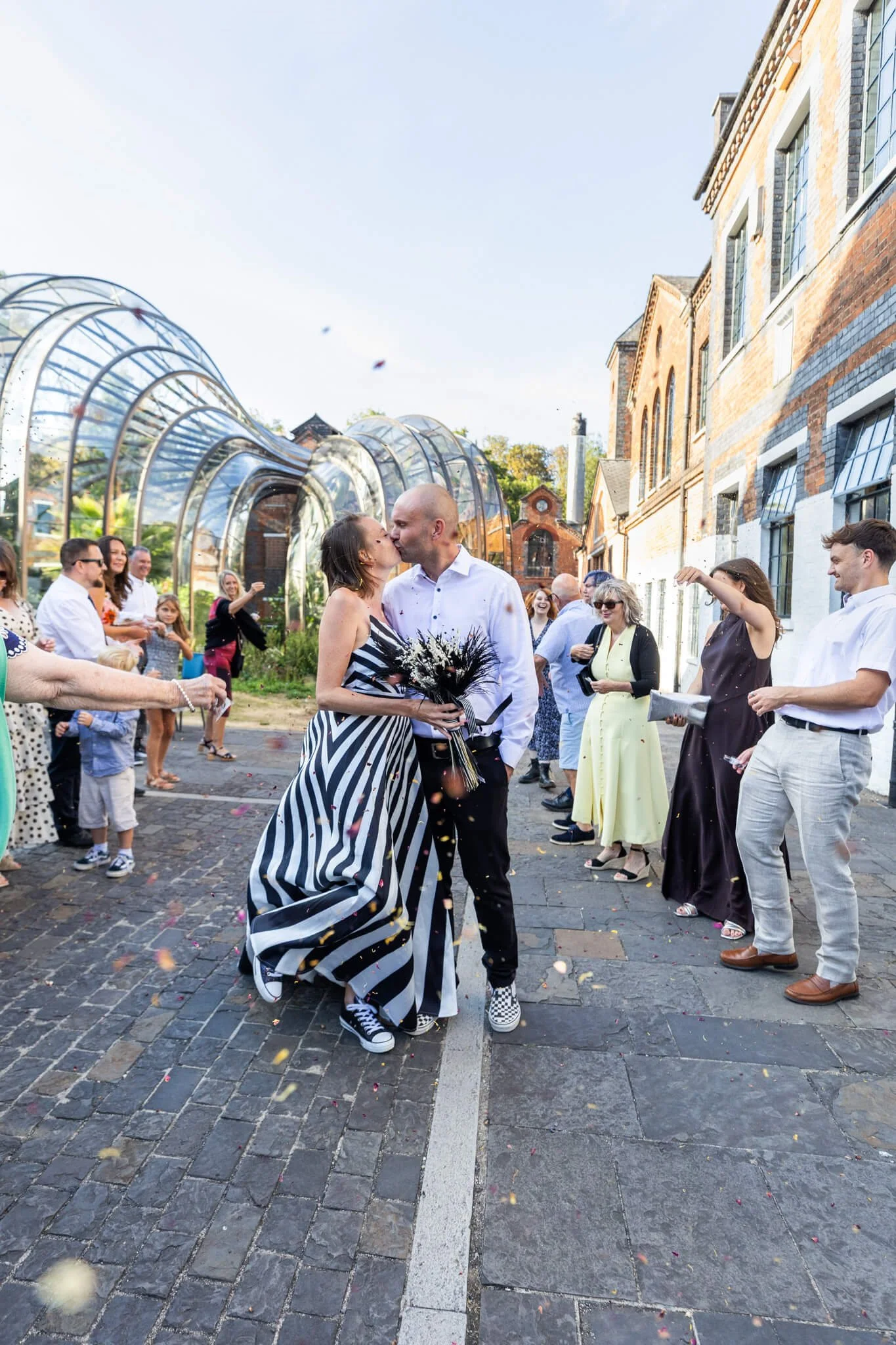 A couple kissing during a wedding celebration outdoors on a cobblestone street. The bride is dressed in a black and white striped dress, and the groom is wearing a white shirt and black pants. They are surrounded by guests, some taking photos and cel