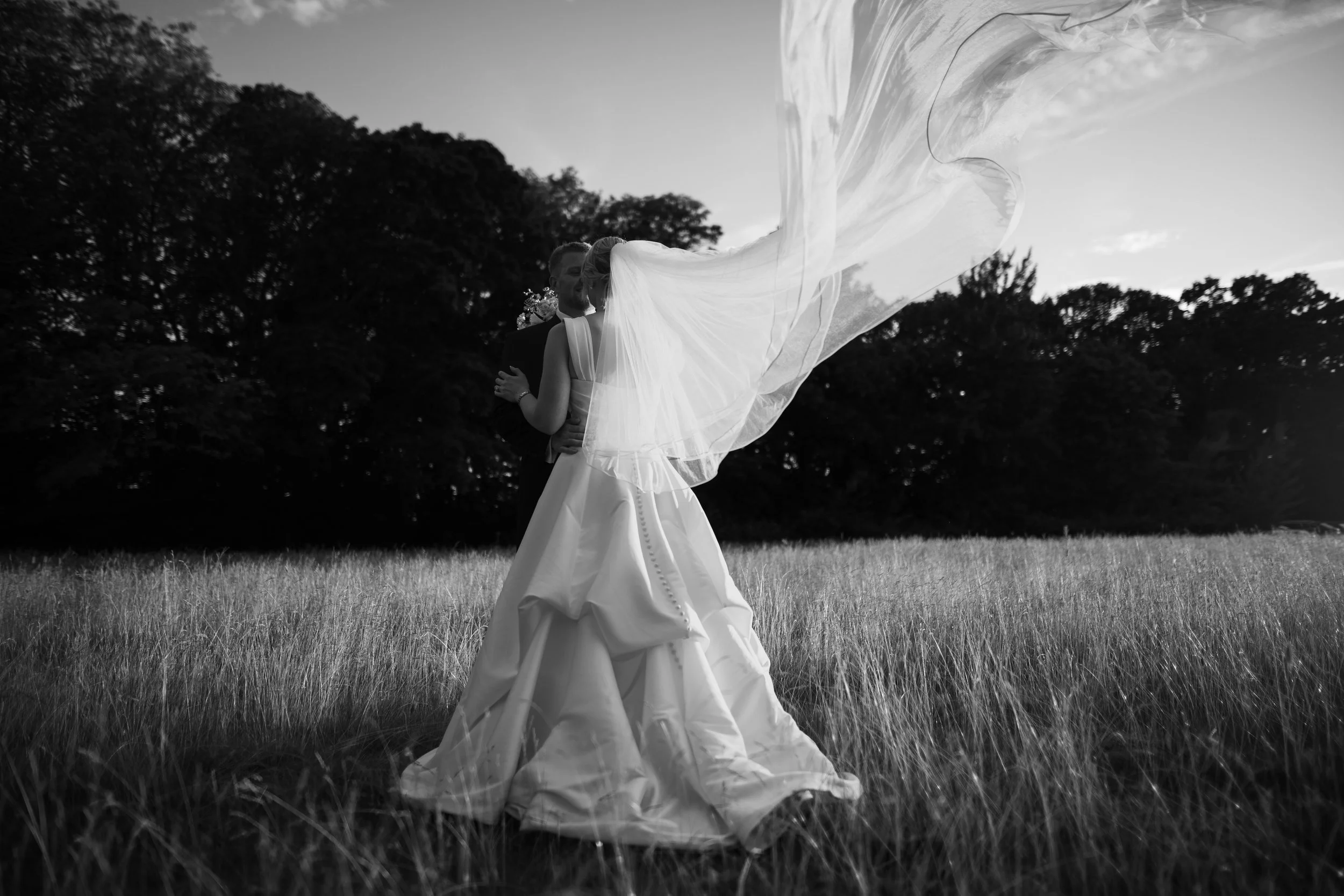 A bride and groom dancing in a grassy field at sunset, with the bride's veil flowing in the wind, captured in black and white.