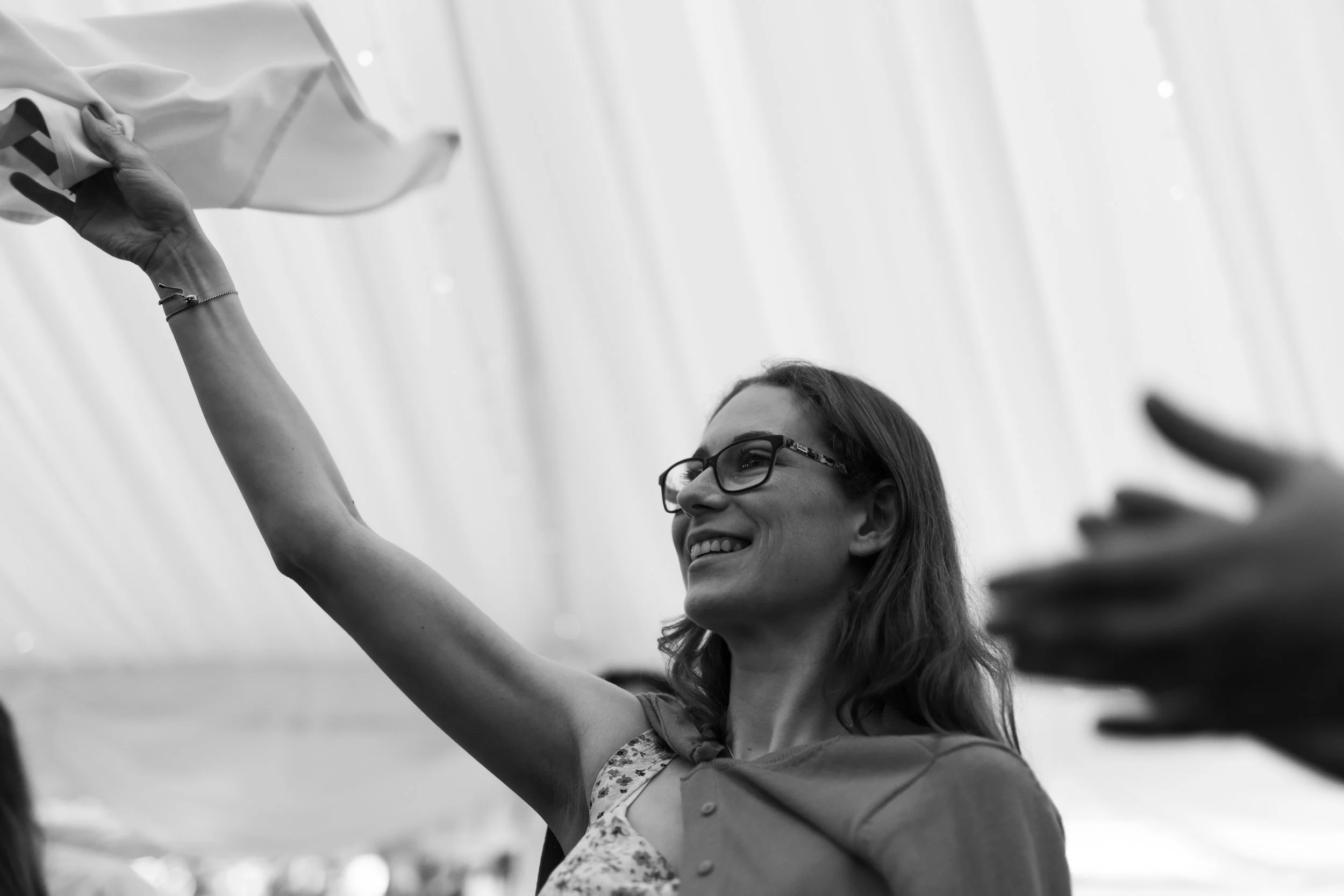 A smiling woman with glasses, raising her arm, holding a fabric, at an outdoor event.