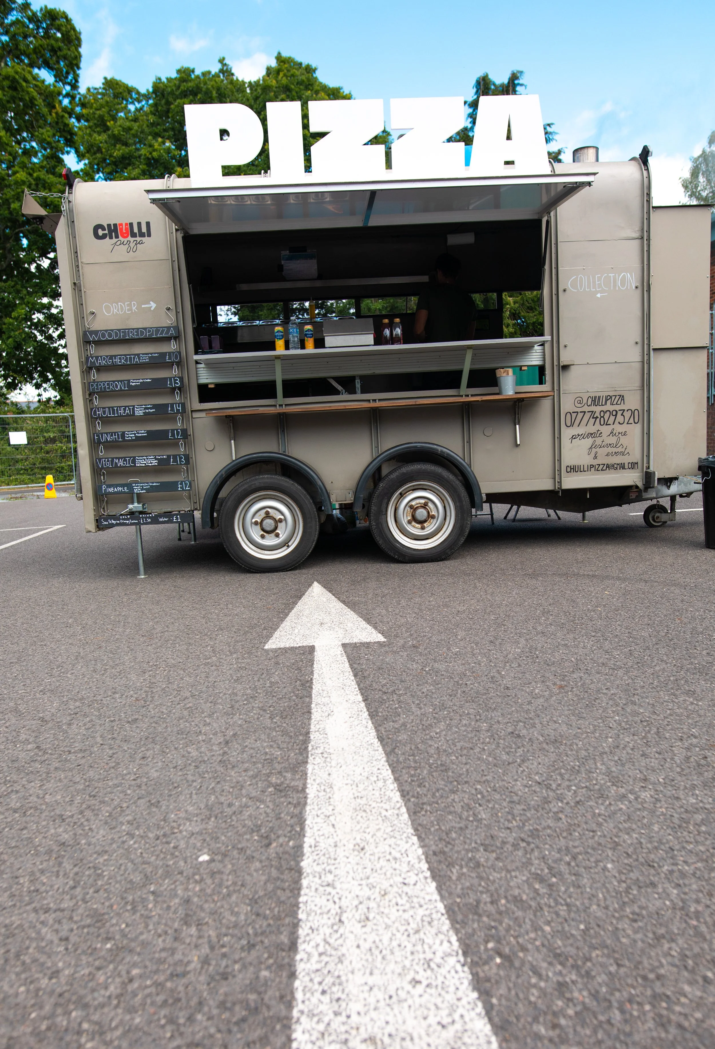 A mobile pizza stand with a large sign reading 'PIZZA' on top, parked in a parking lot with greenery and trees in the background. A white arrow painted on the ground points toward the stand.