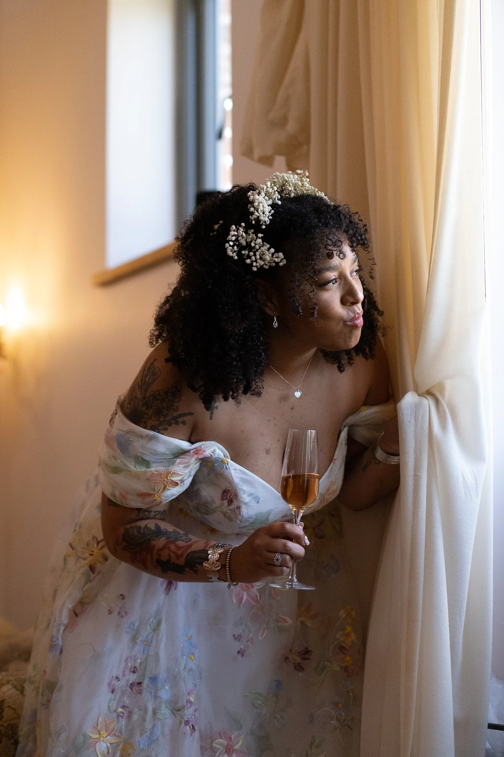 A woman with curly hair and floral decorations wearing a floral off-the-shoulder dress, holding a glass of wine, looking out the window.