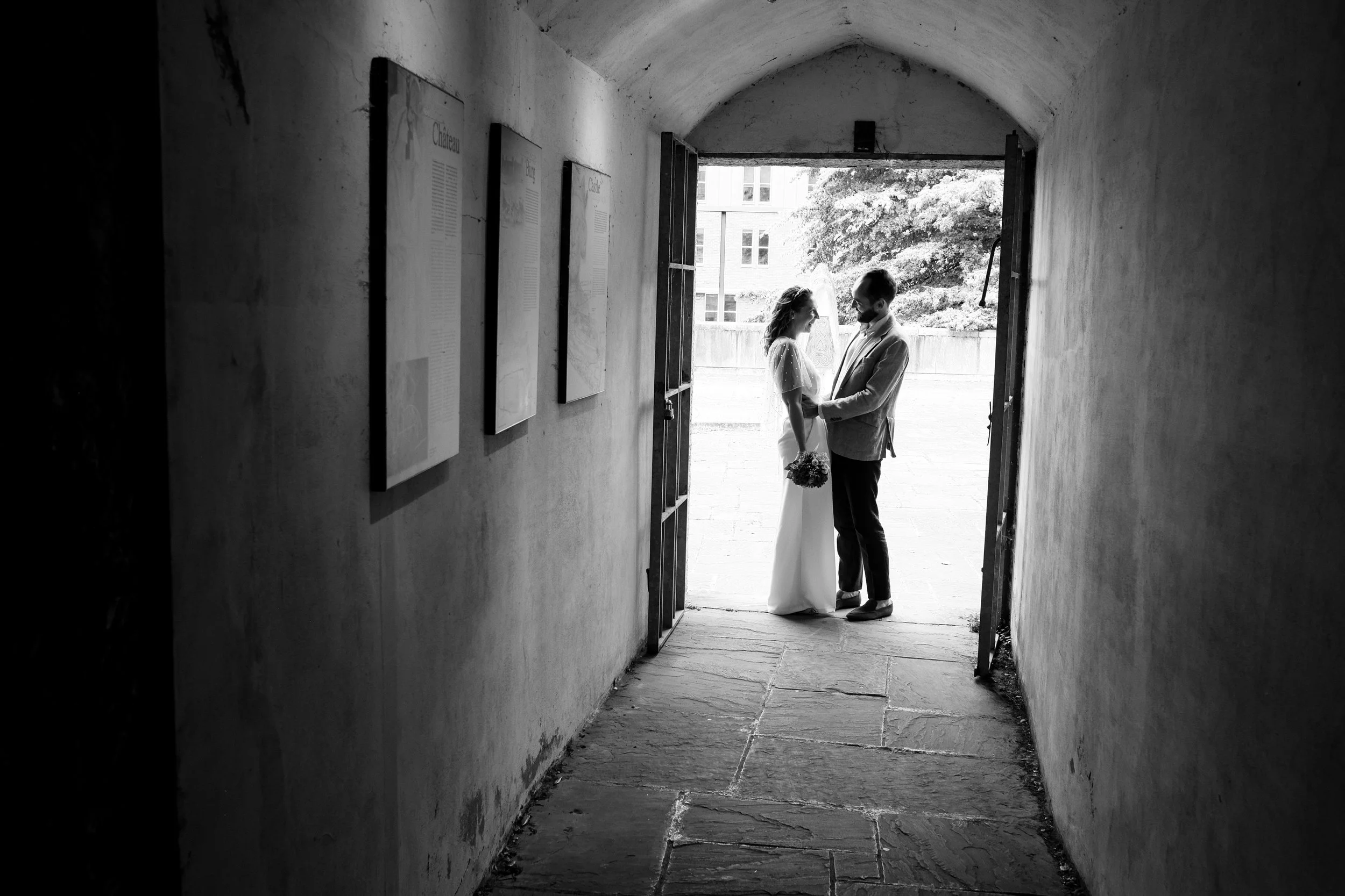 A black and white photo of a couple standing close together in a tunnel-like space, holding hands and smiling at each other. The scene is lit from outside, with a window and tree visible in the background.