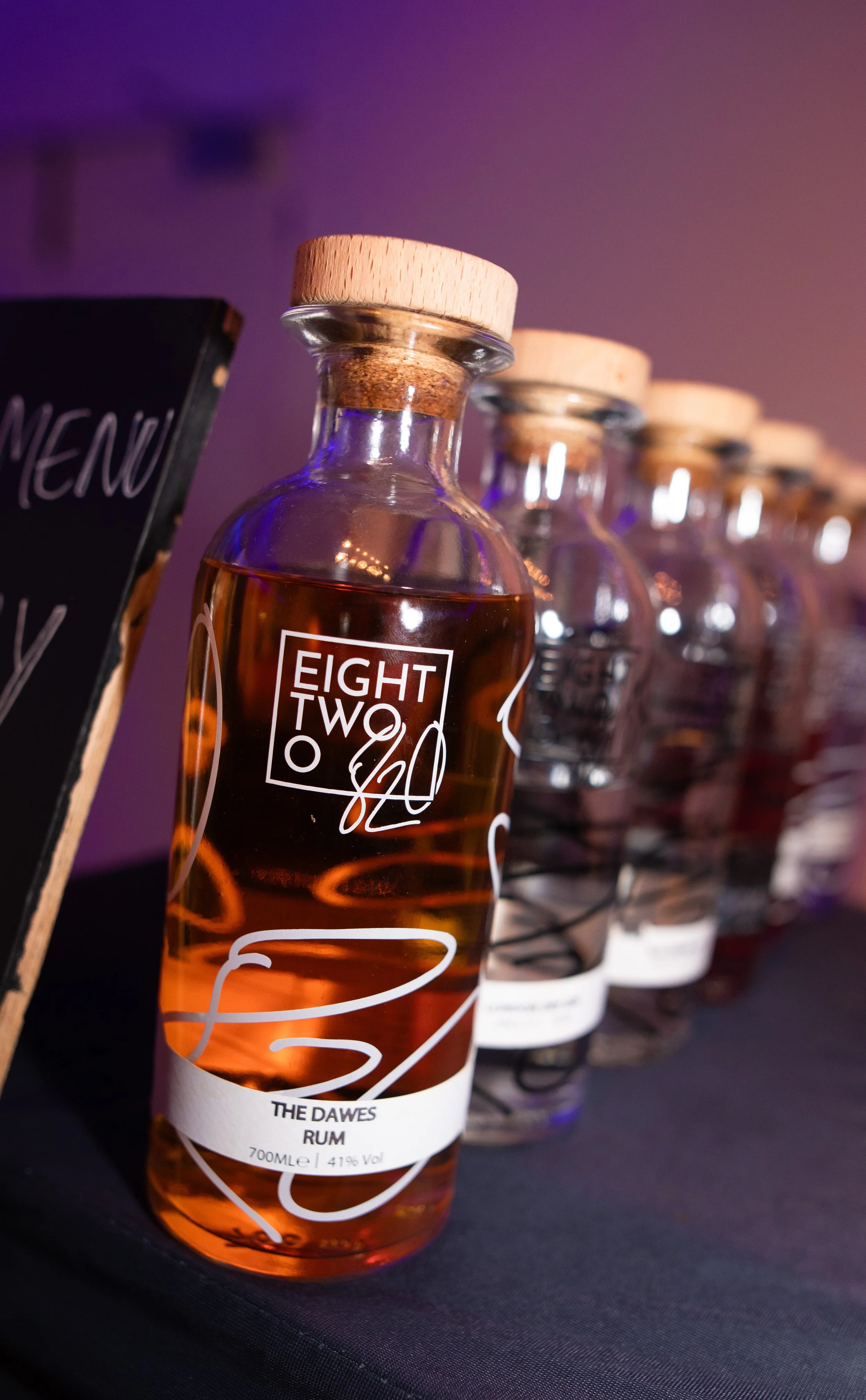 Line of glass bottles with wooden caps, the one in front labeled "The Dawes Rum," containing amber-colored liquid, on a dark surface with a purple background.