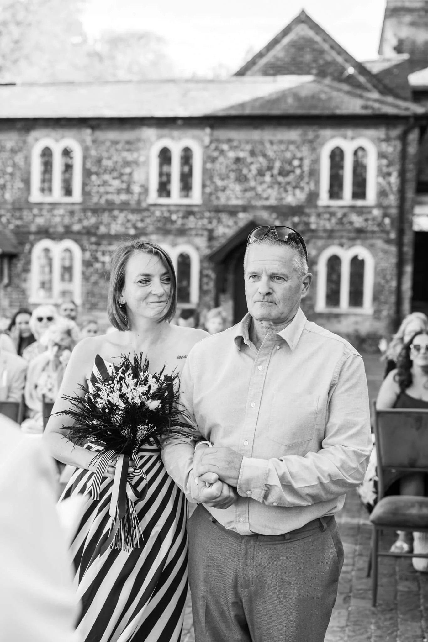 A woman and a man are standing close together outdoors, seemingly at a formal or community event. The woman is holding a bouquet of flowers and is wearing a striped dress with a floral corsage, while the man is dressed in a light-colored button-up sh