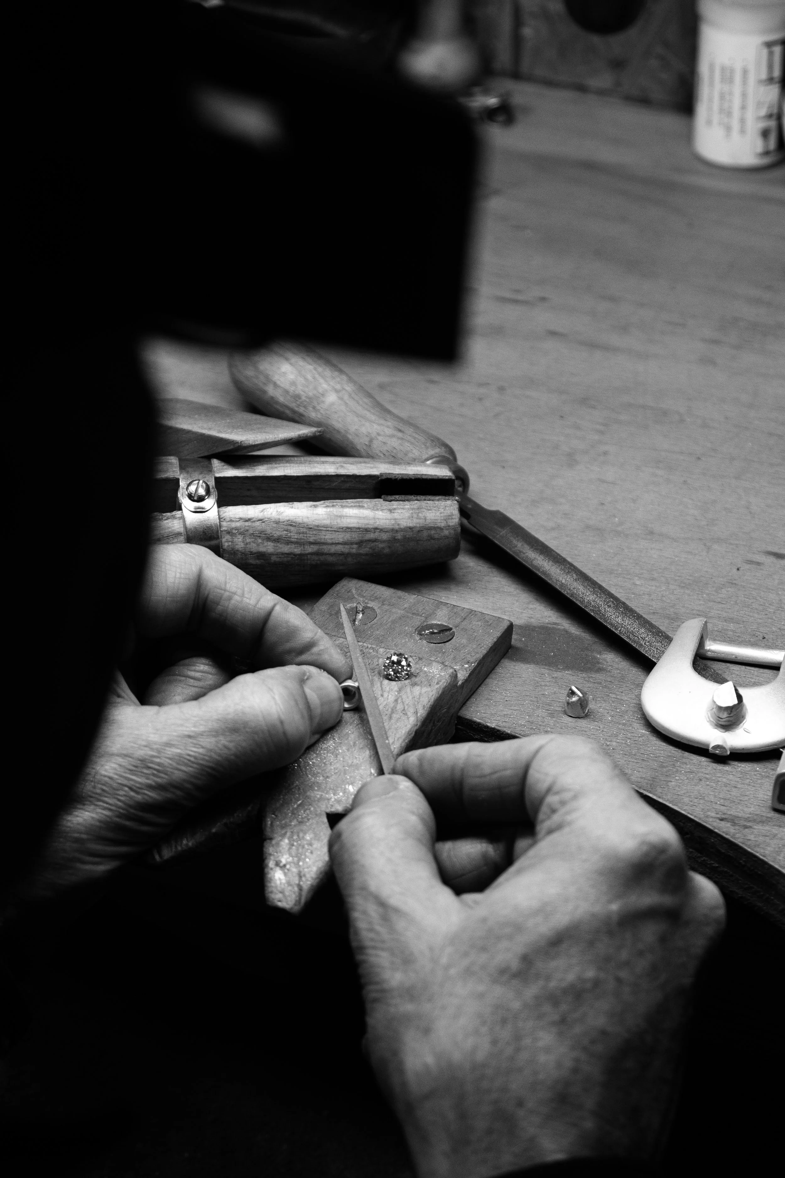 Close-up black and white photo of hands working on a jewelry piece, with various tools and jewelry pieces on a wooden work surface.