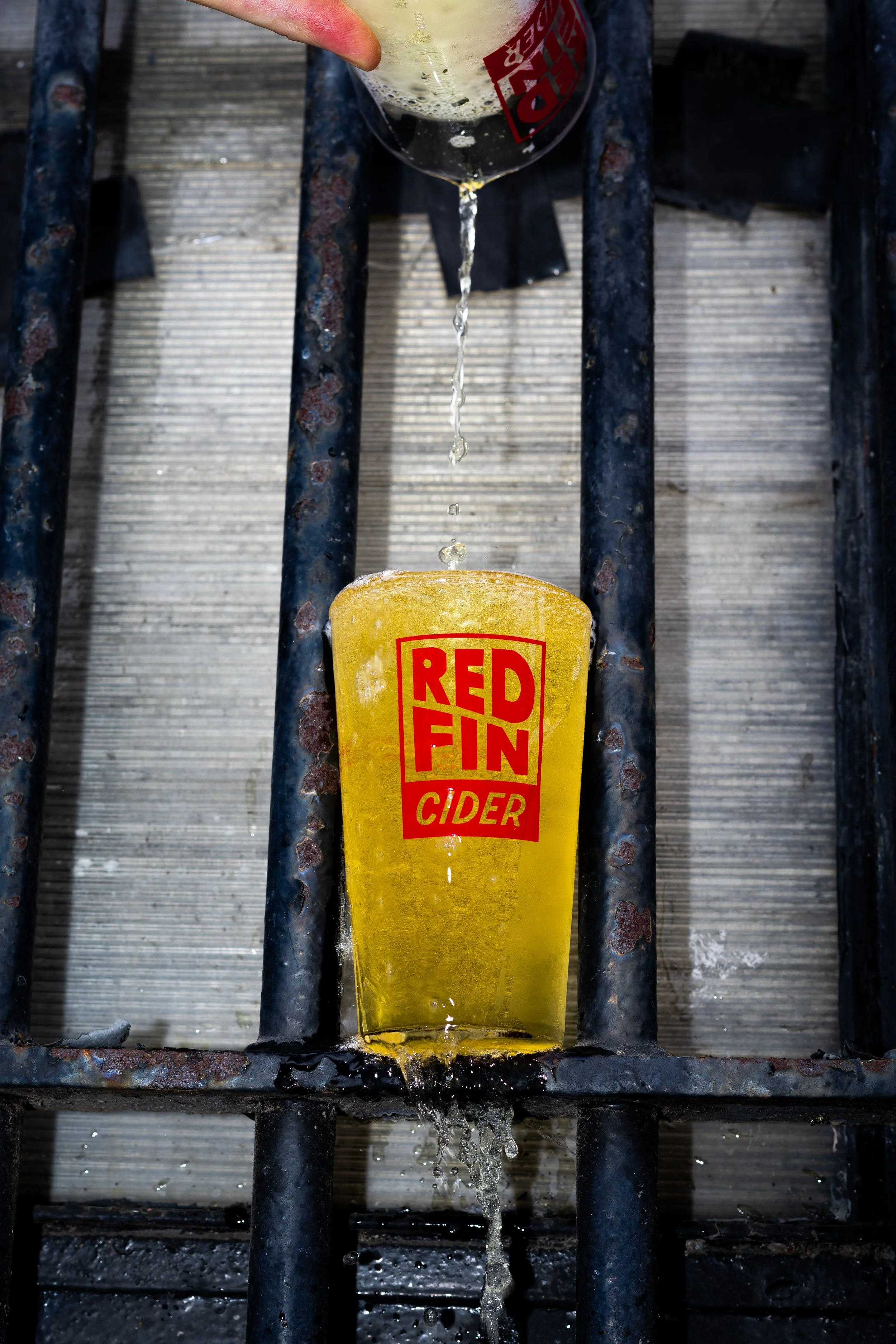 Yellow cup with red text 'Redfin Cider' underneath a metal grate, with liquid pouring into the cup and spilling over the edge.