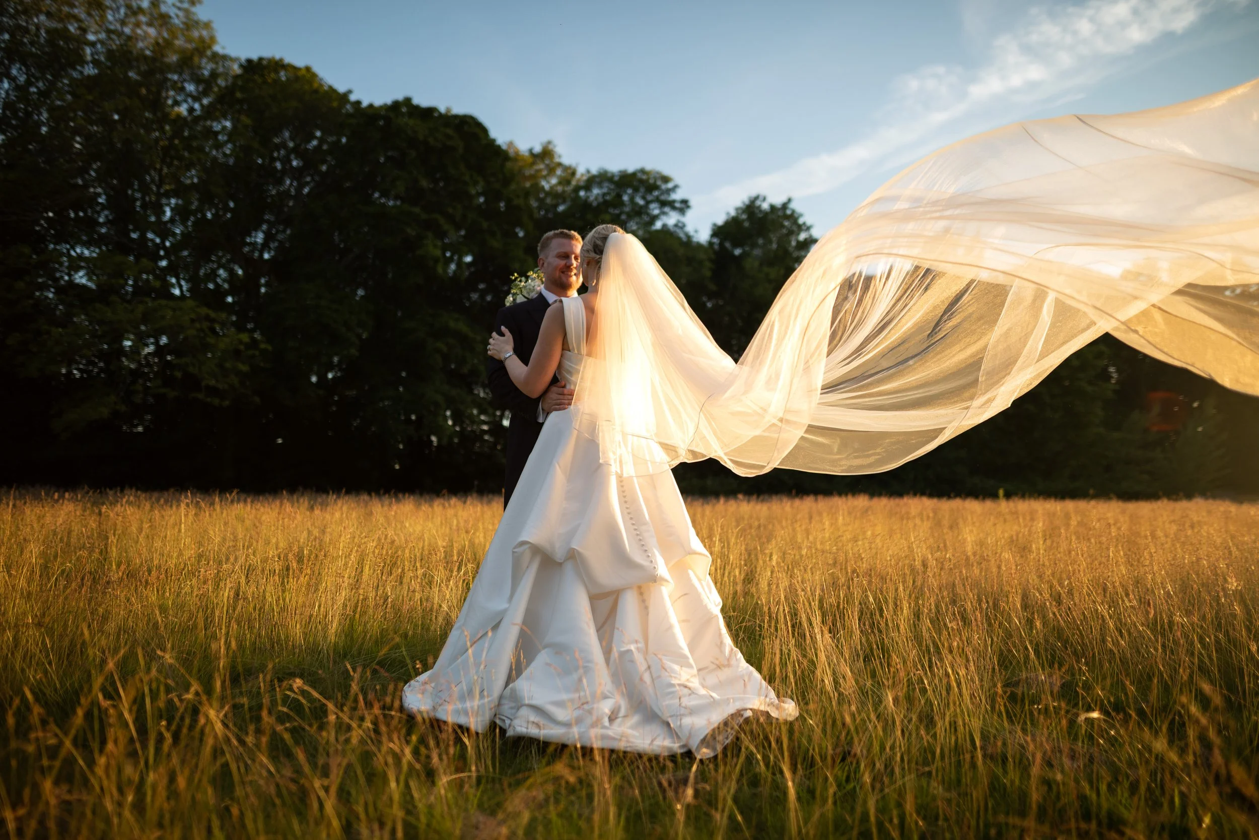A bride and groom sharing a moment outdoors during sunset, with the bride wearing a flowing white gown and veil, and the groom in a dark suit, standing on a grassy field with trees in the background.