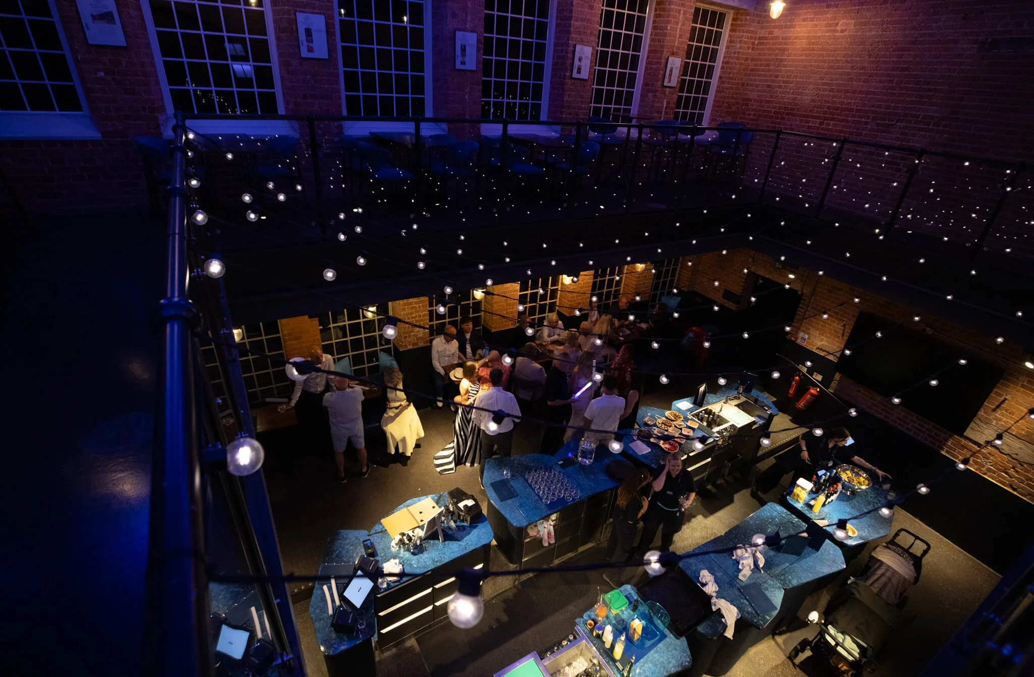 Indoor party scene with people gathered around tables with food, bar area, and string lights, in a brick-walled venue.