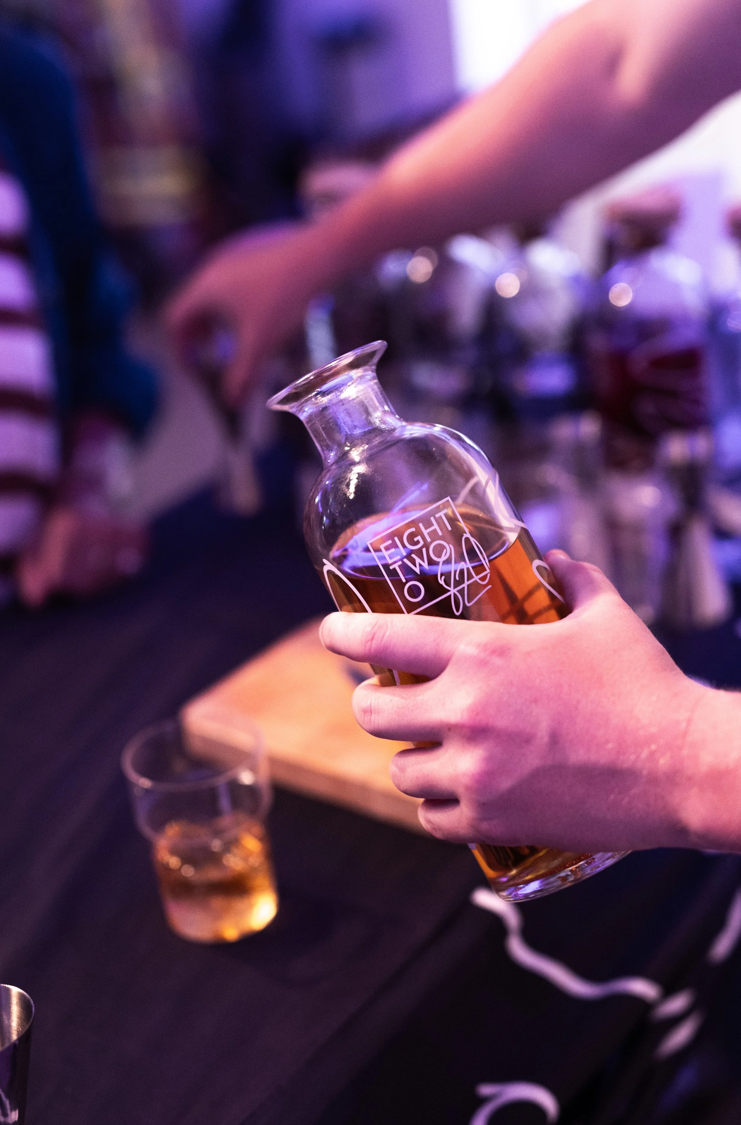 A person holding a glass bottle of amber-colored beverage labeled 'EIGHT TWO' with a signature, in front of a table with a shot glass filled with the same beverage and a blurred row of bottles in the background.