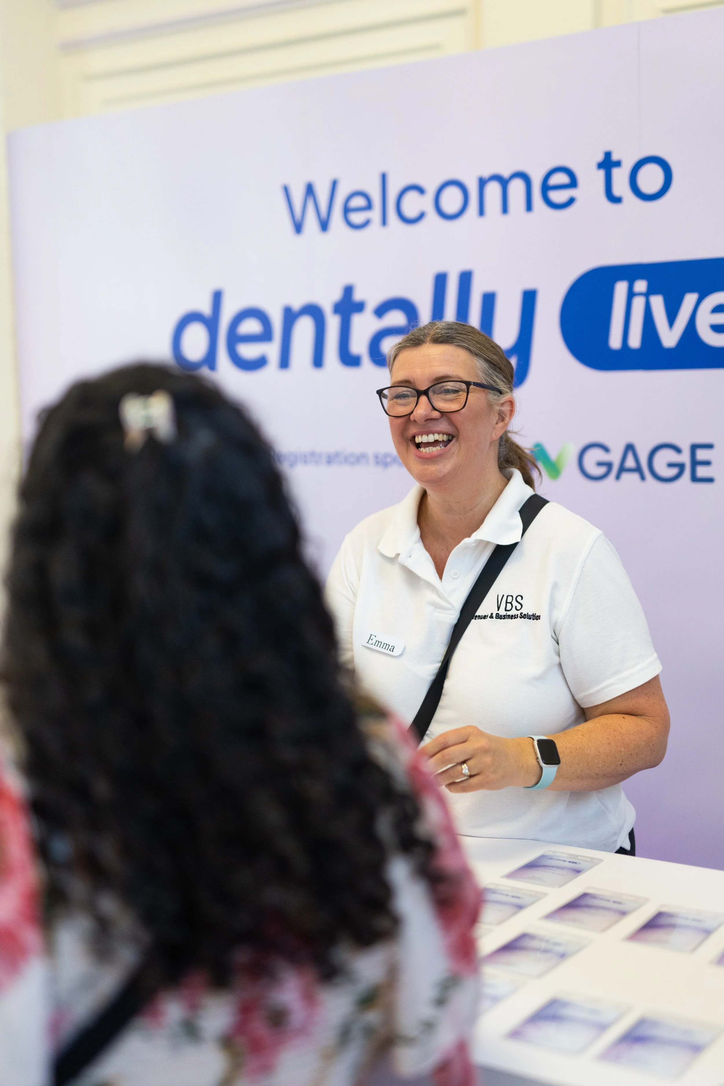 A smiling woman with glasses and a white shirt standing in front of a large sign that reads 'Welcome to dentally live'. She is facing a person with curly dark hair and a floral top, who is out of focus in the foreground. There are brochures or papers