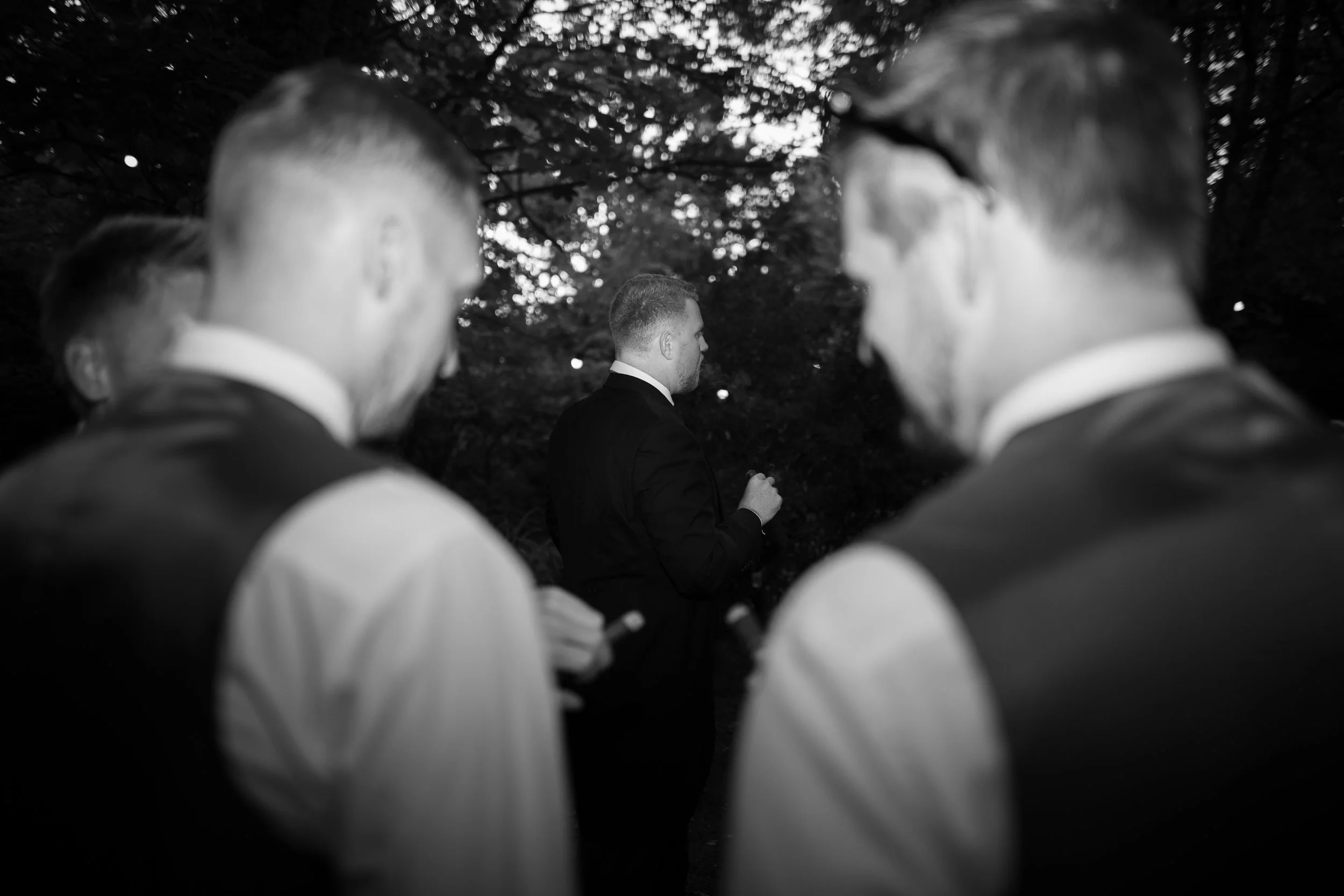 Black and white photo of three men in suits, two of whom are blurred, facing a man with short hair in a suit, outdoors with trees in the background.