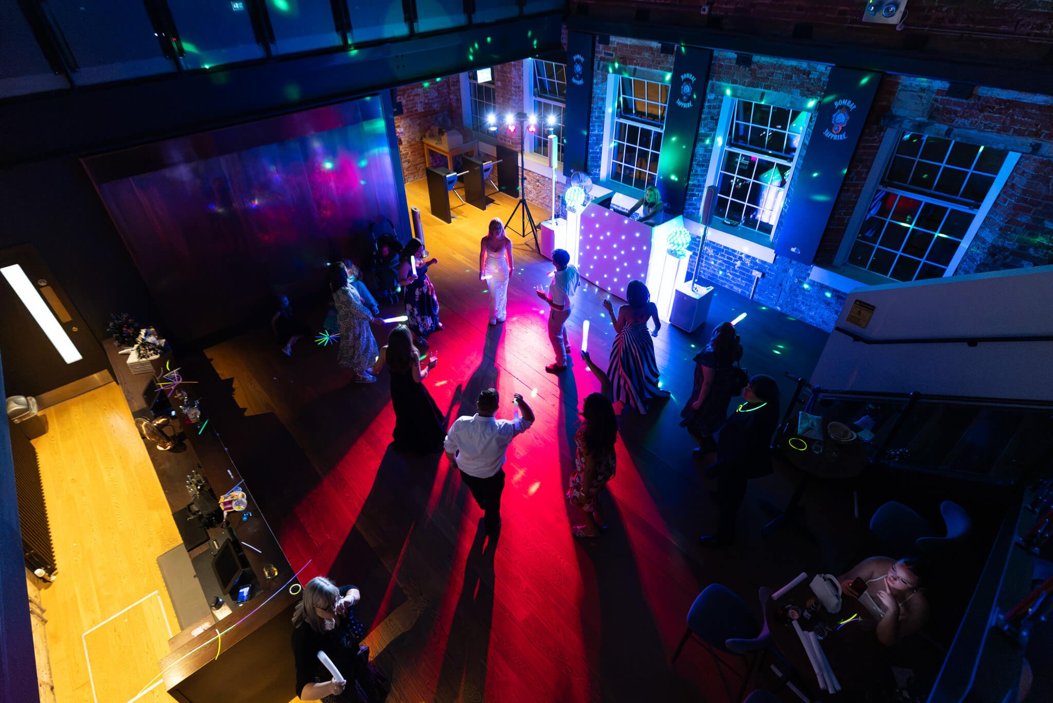 People dancing on a wooden floor illuminated with colorful lights, in a room with exposed brick walls and large windows, during a nighttime party or celebration.