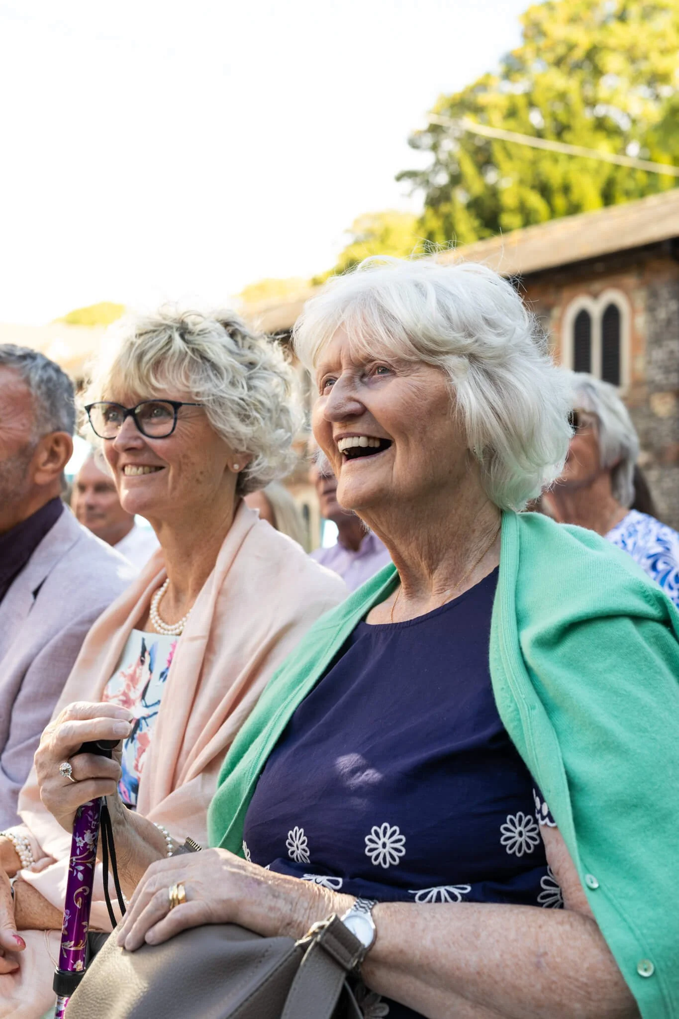 A group of elderly people sitting outdoors during daytime, smiling and enjoying an event. The background shows trees and part of a brick building.