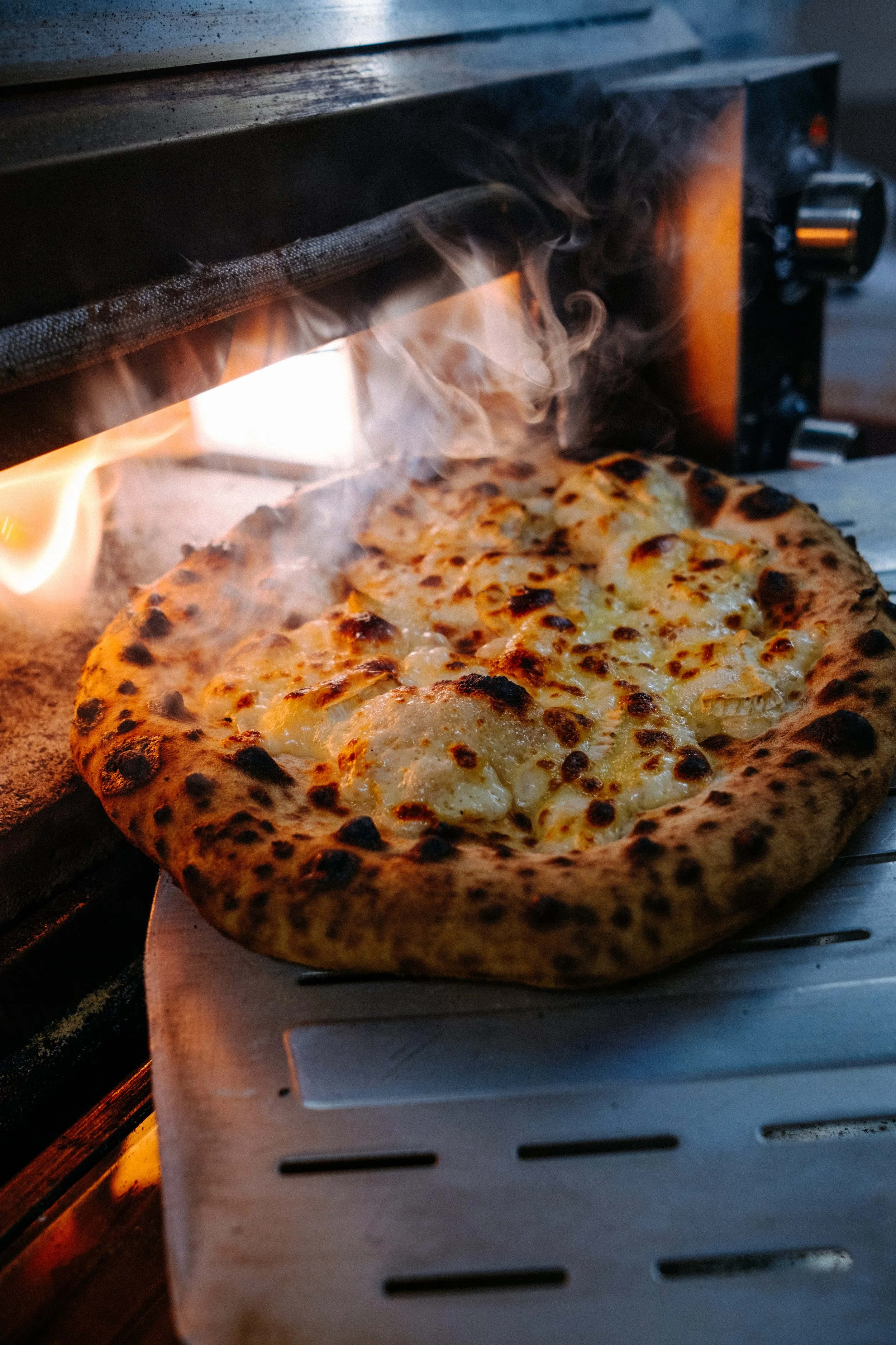 Close-up of a cheese pizza baking in a wood-fired oven with flames visible and smoke rising.