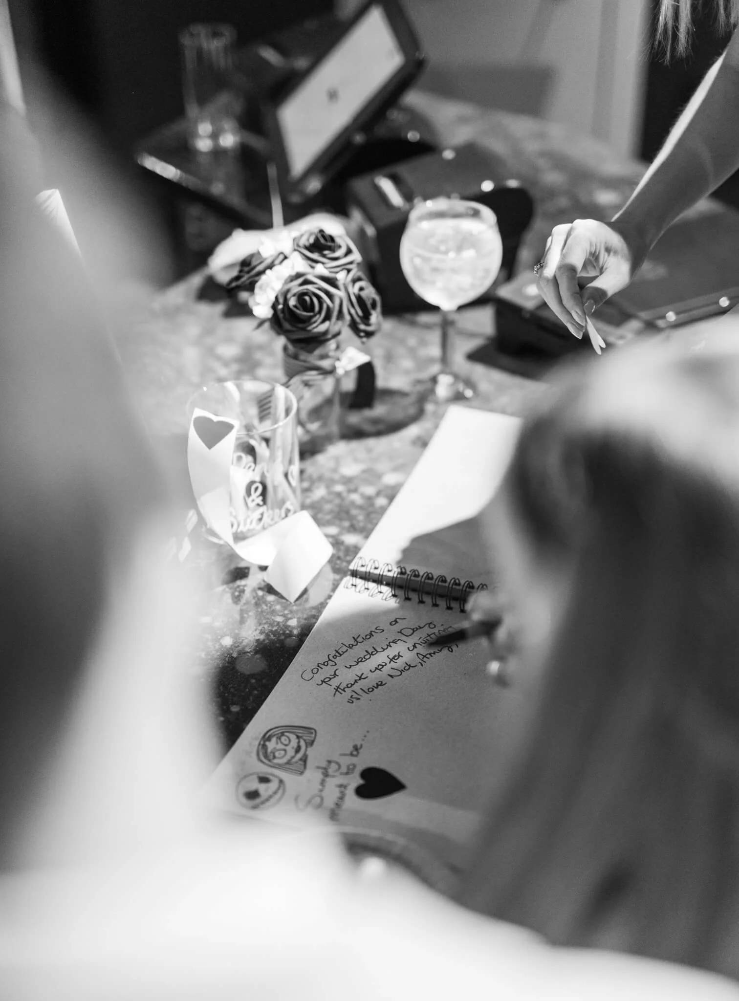 A handwritten thank-you note on a lined piece of paper, with heart and smiley face stickers, surrounded by a few decorative objects including a bouquet of roses, a glass of water, and some electronic devices on a countertop.