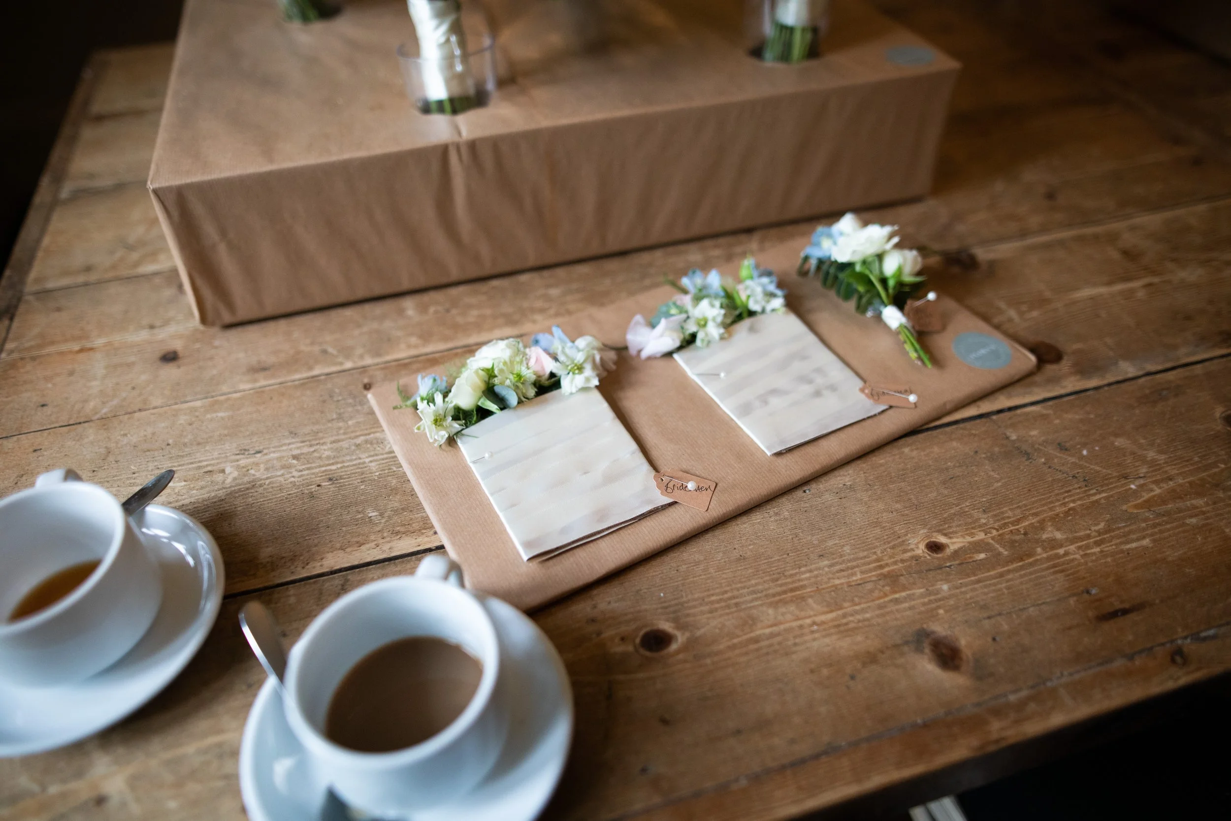 A wooden table with four white cups of coffee, and a decorative floral arrangement on small standing cards labeled "bride" and "groom".
