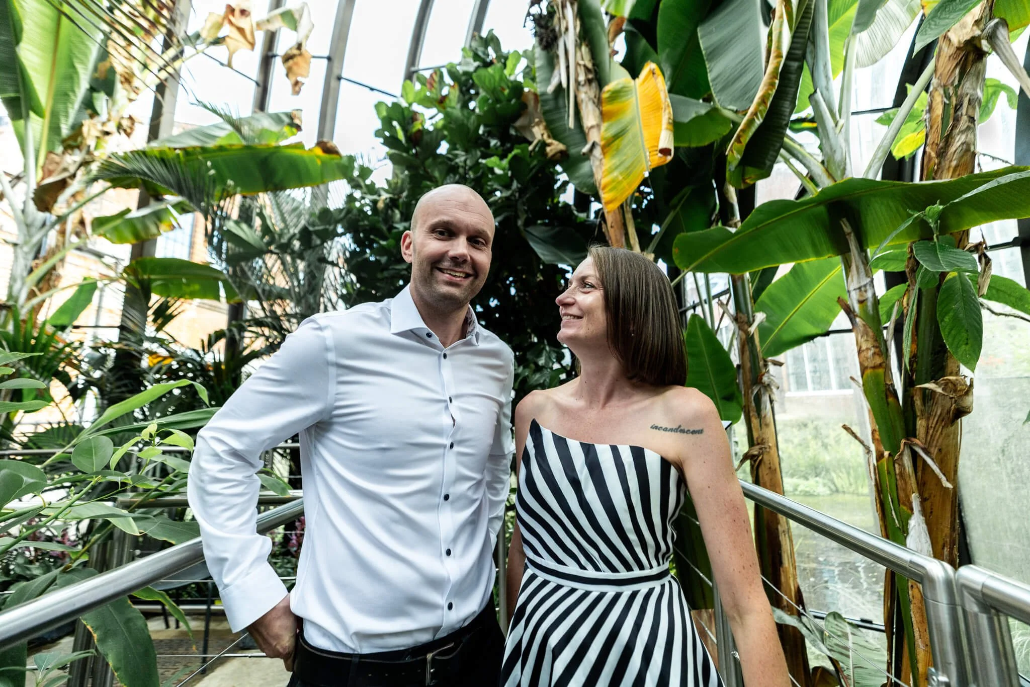 A man and woman standing together in a greenhouse filled with large green tropical plants. The man is wearing a white dress shirt, and the woman is dressed in a black and white striped sleeveless dress with a tattoo on her chest. They are smiling and