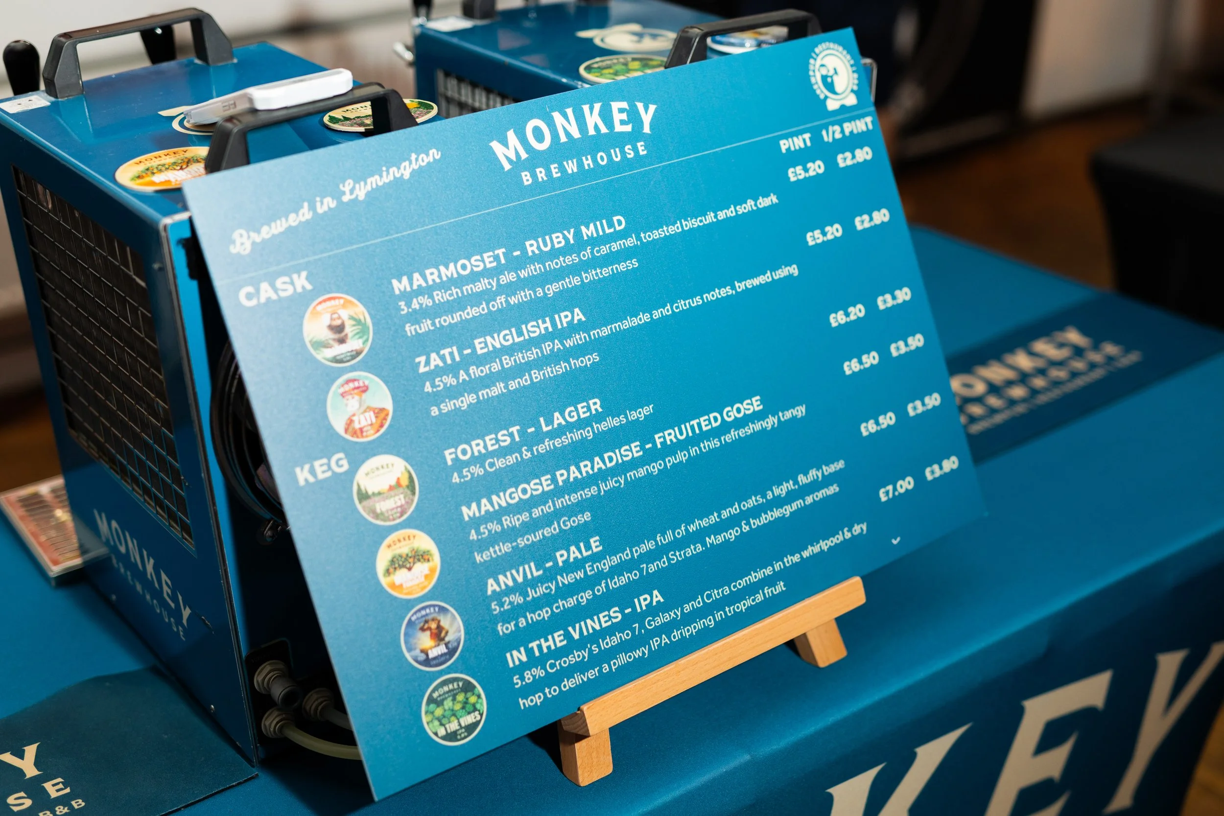 Close-up of a blue beer menu board on a small wooden stand at a brewery, listing various beers and their descriptions and prices.