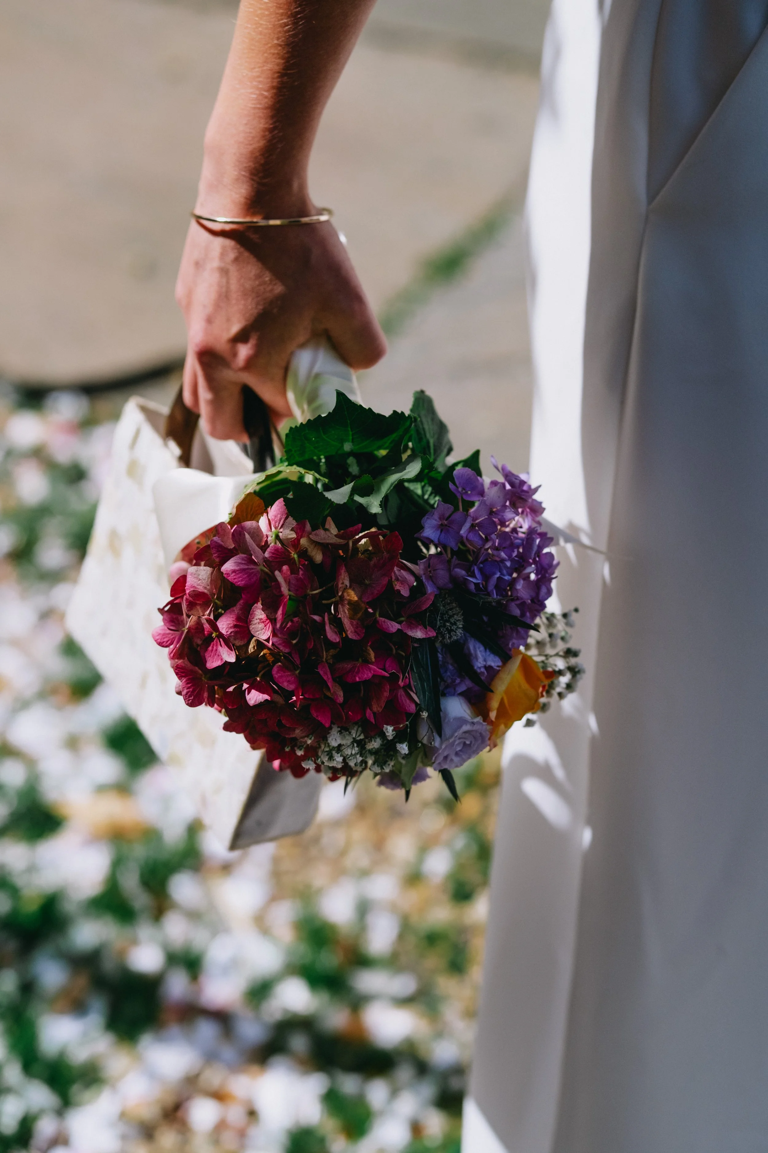 A person holding a bouquet of pink, purple, and yellow flowers, wearing a gold bracelet and a white outfit, standing outdoors in sunlight.