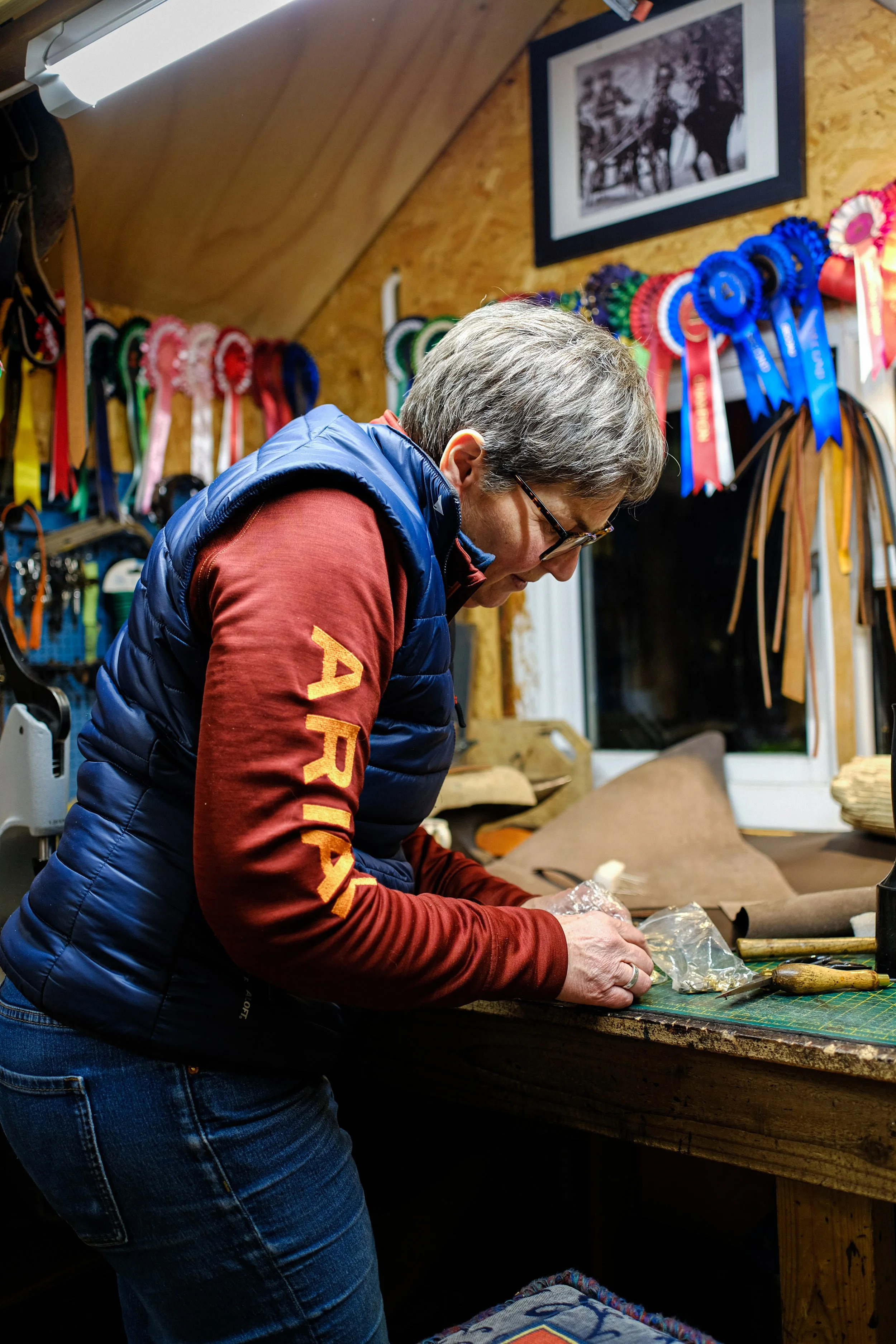 A person with short gray hair and glasses working at a cluttered wooden workbench, surrounded by colorful ribbons hanging on the wall and framed black and white photograph, in a woodworking or craft shop.