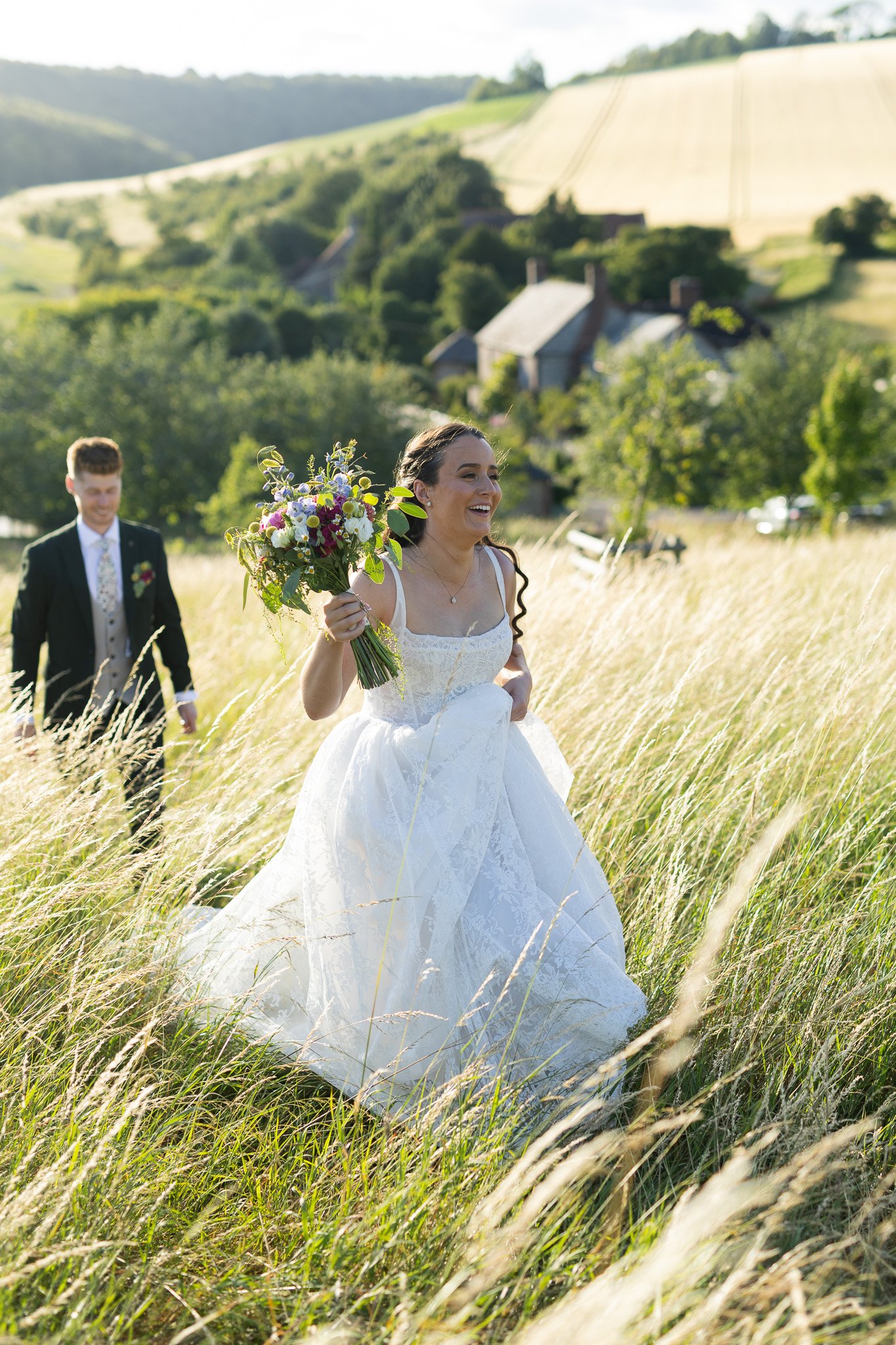 A bride walks ahead of the groom carrying her flowers  in the sun drenched meadow at upwaltham barns