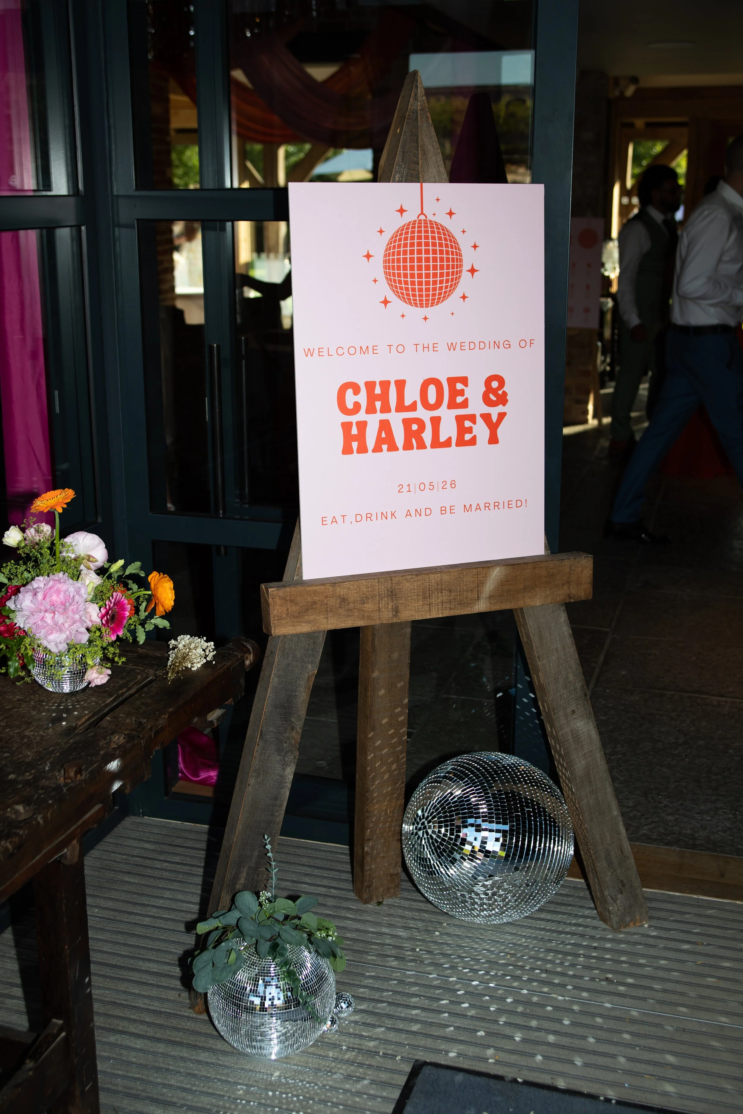 A wedding welcome sign for Chloe and Harley with a disco ball design, decorated with flowers and placed on a wooden stand inside a venue.