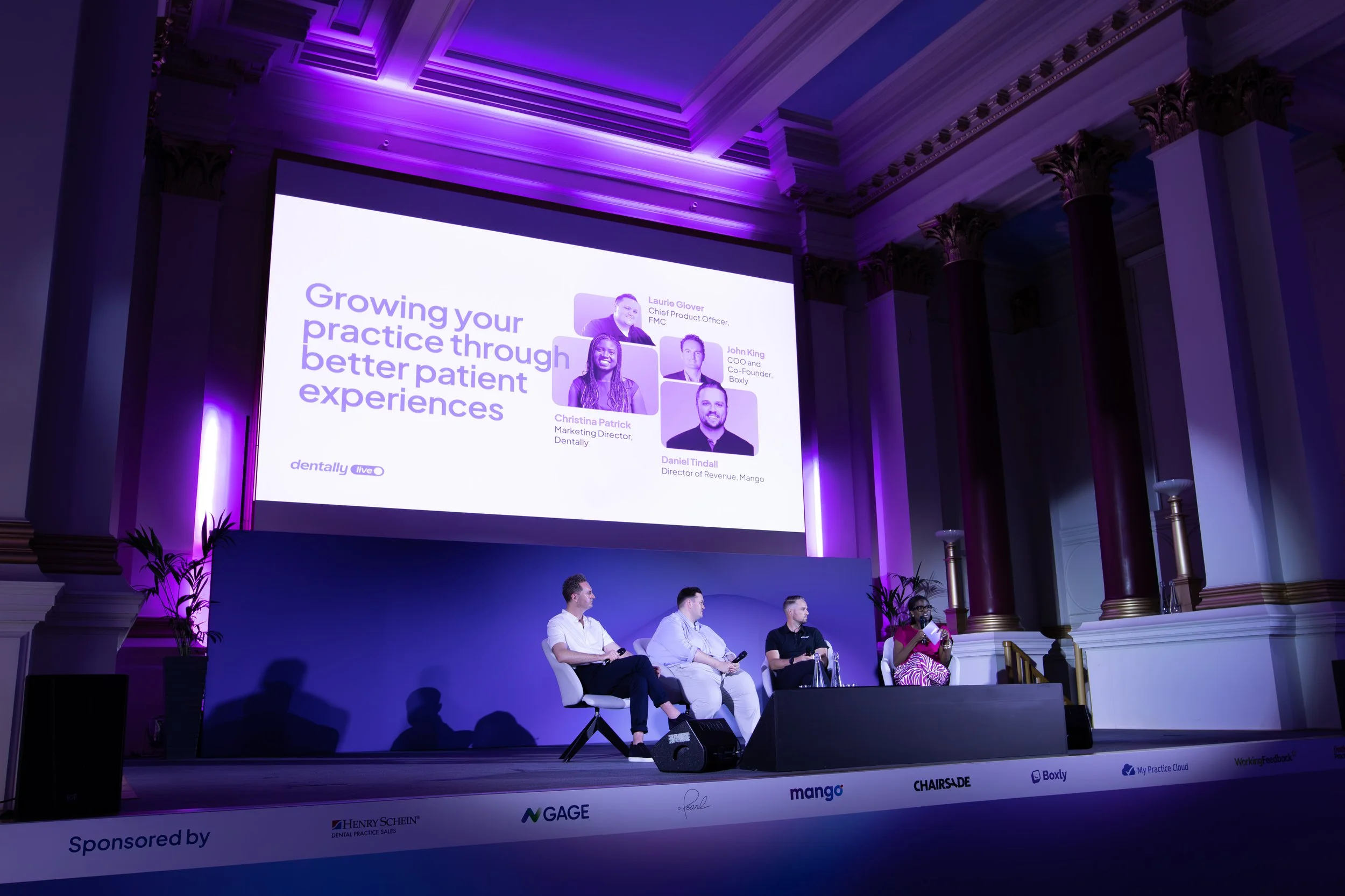 A panel of five people seated on stage during a conference or presentation in a room with ornate columns and purple lighting. A large screen behind them displays the title 'Growing your practice through better patient experiences' along with photos a