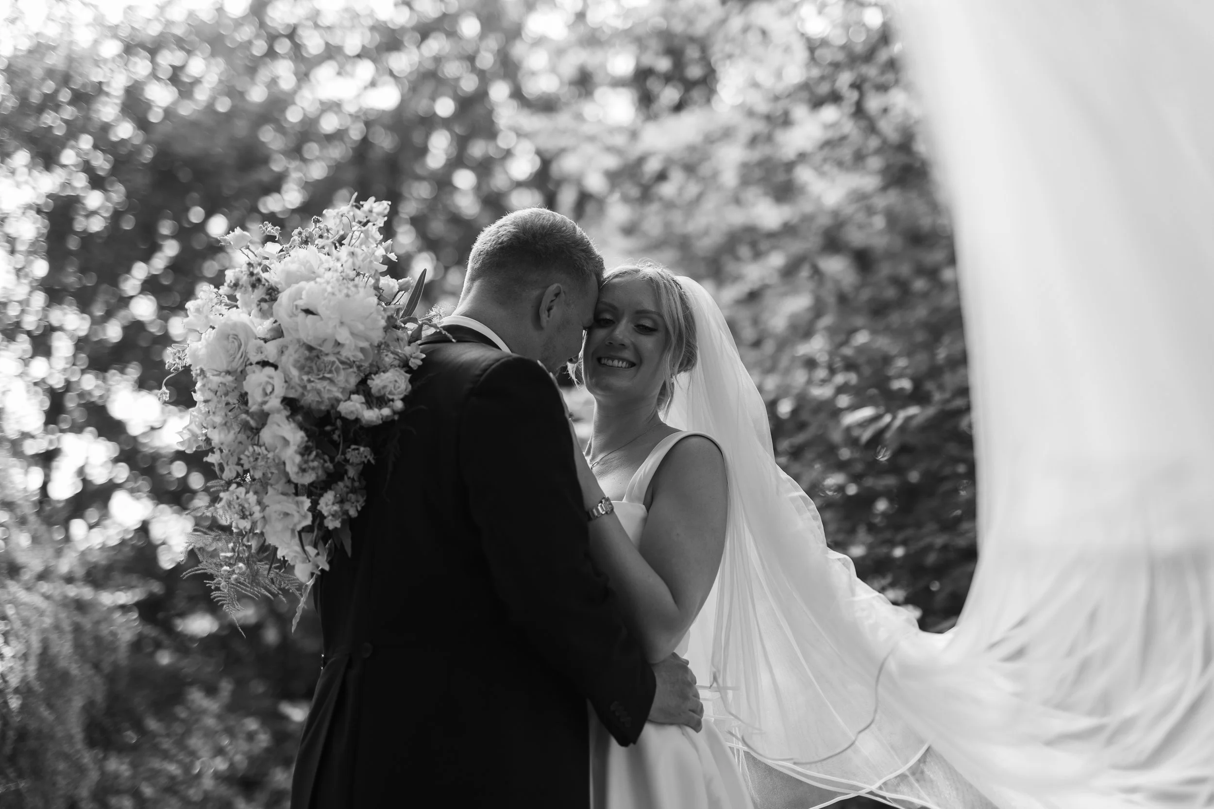 A black and white photo of a bride and groom touching foreheads during their wedding, with the groom holding a large bouquet of flowers.