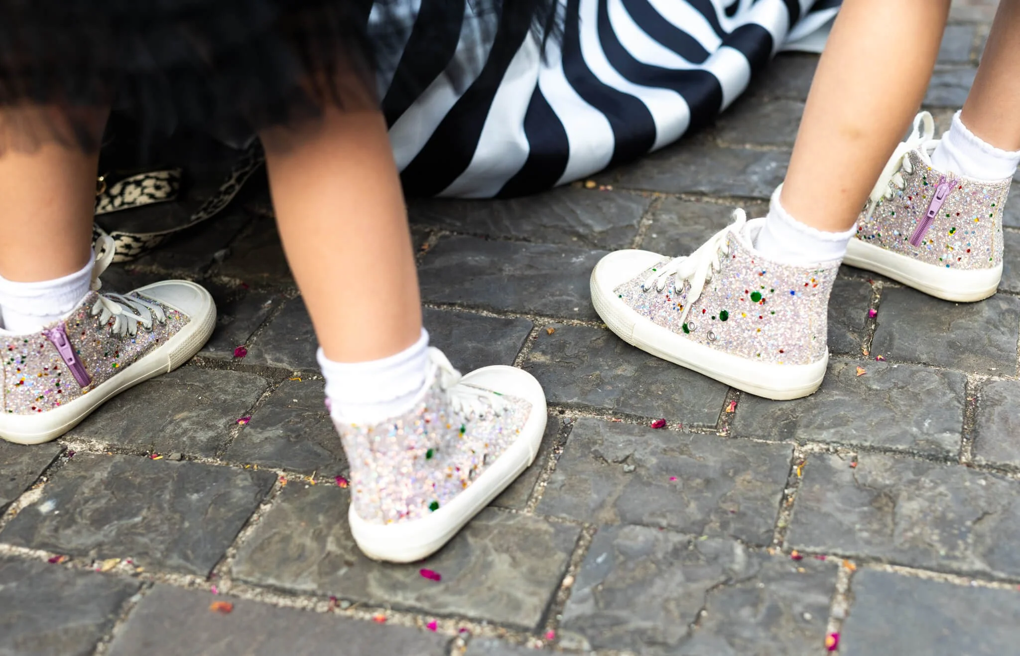 Close-up of children's glittery, decorated sneakers on a cobblestone street, with some scattered confetti.