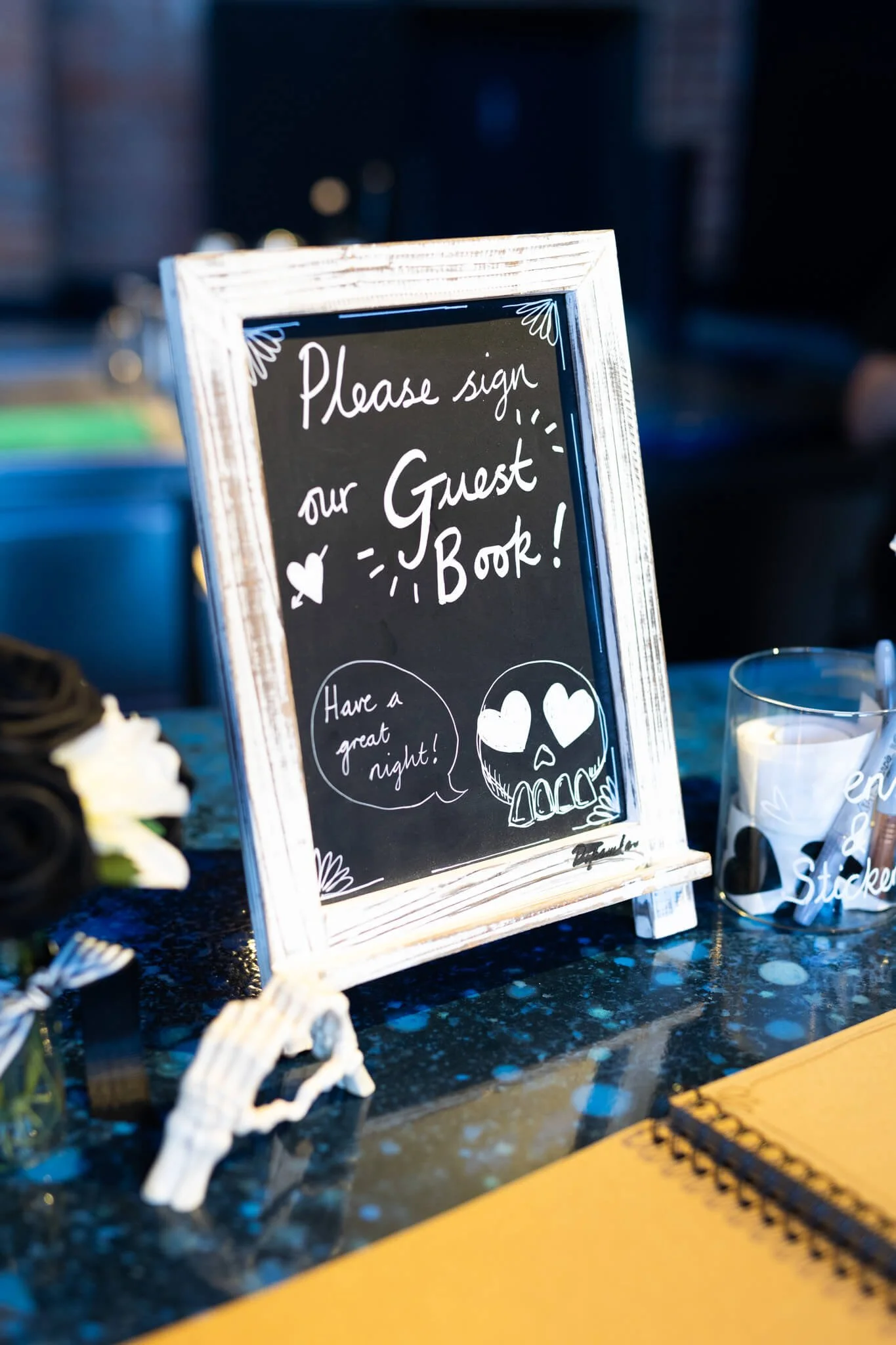 A chalkboard sign with a wooden frame displays a message that says, 'Please sign our Guest Book!' with decorative drawings, including a skull with hearts in its eyes and speech bubbles, placed on a dark speckled countertop with items like a skeleton 