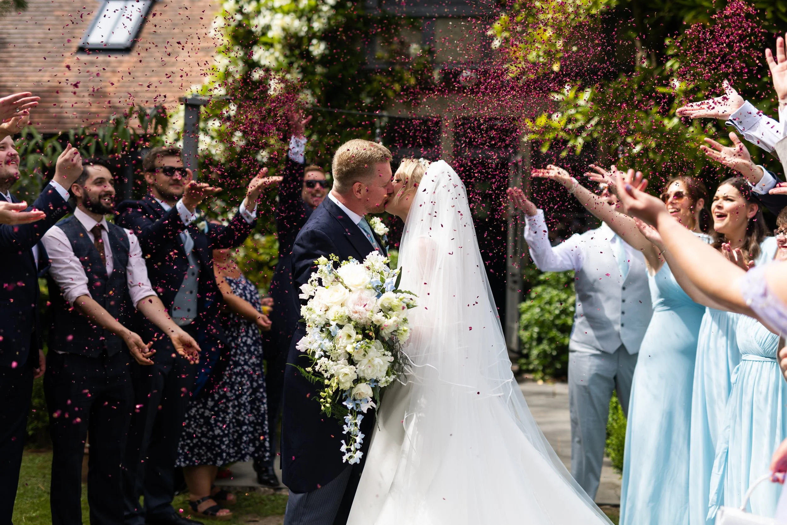 A bride and groom kissing during their wedding celebration amid friends throwing pink and purple flower petals in the air on a sunny day, outdoors with greenery and house in the background.
