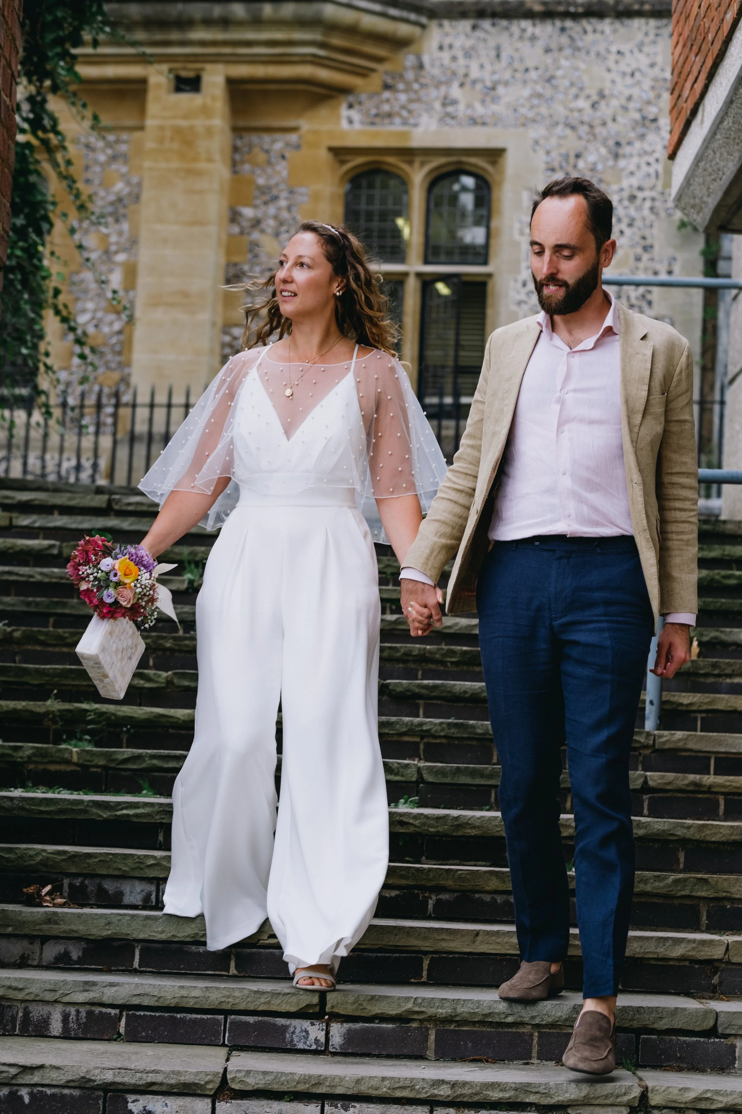 A bride and groom walking hand in hand down stone steps outside a historic building with brick and stone walls, large arched windows, and greenery.