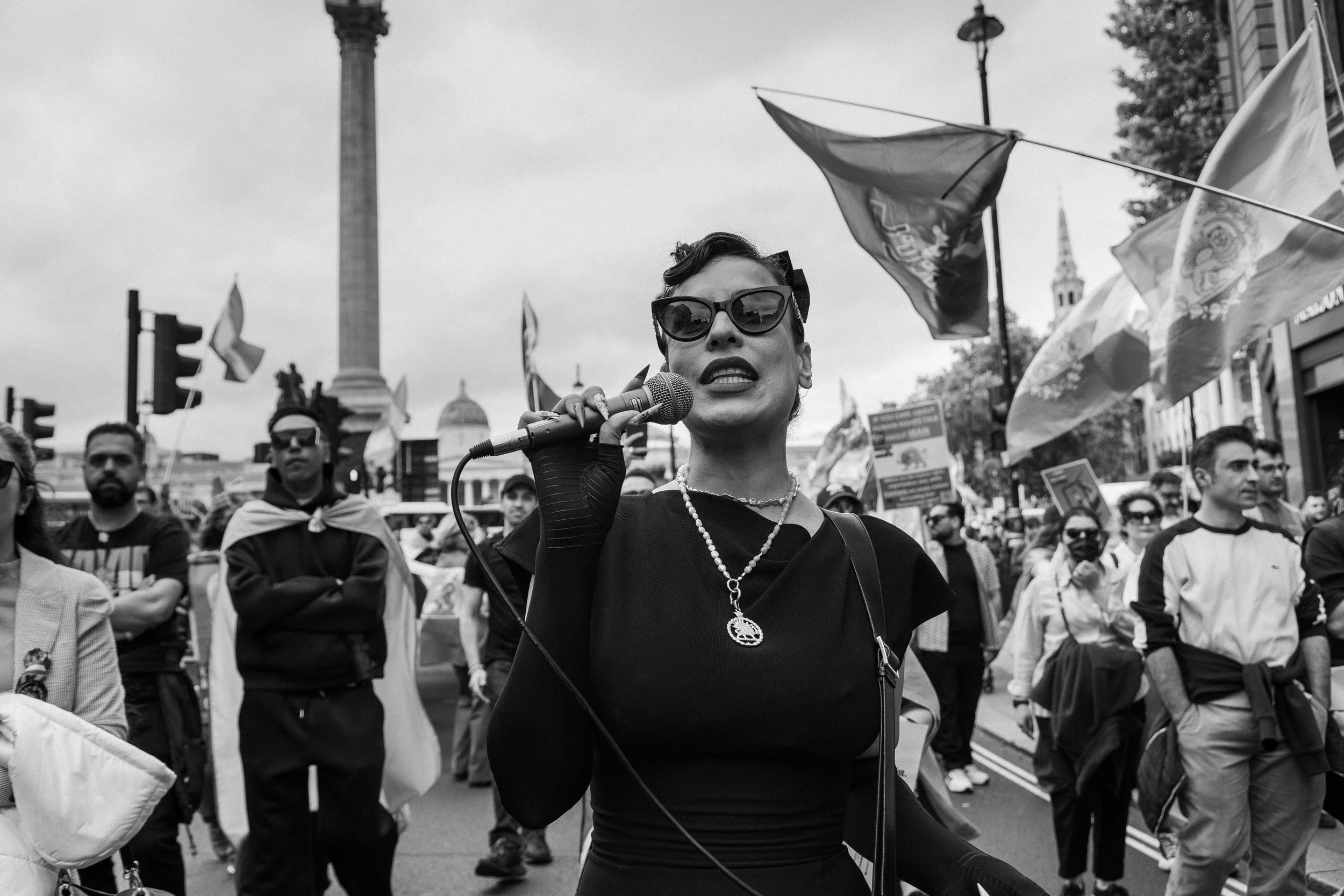 A woman wearing sunglasses, headphones, and a necklace speaking into a microphone at a protest or rally, with a crowd and flags in the background in an urban setting.