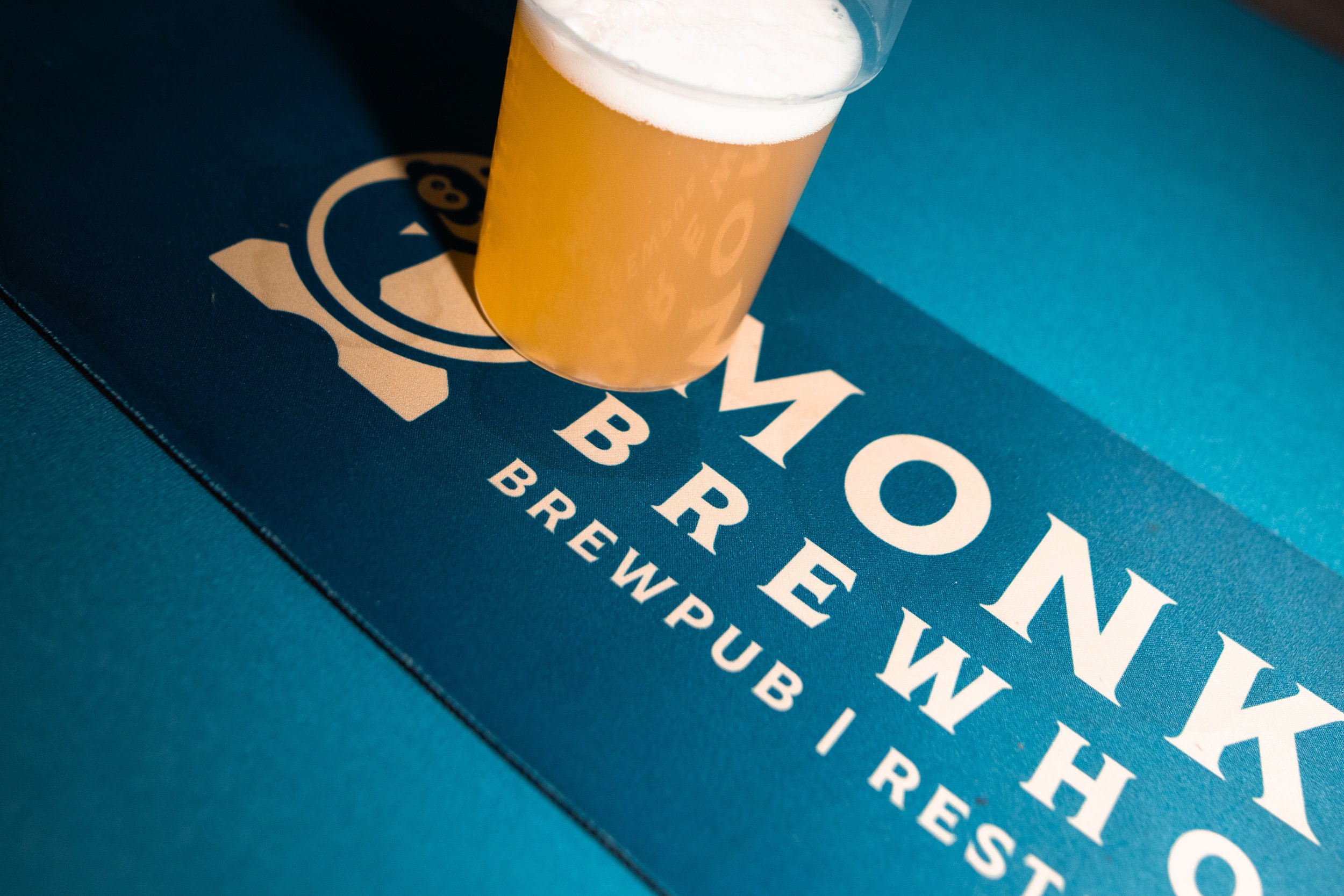A pint glass filled with a hazy, light-colored beer with a frothy white head, placed on a blue surface with the partially visible words and logo of a brewery or beverage brand.