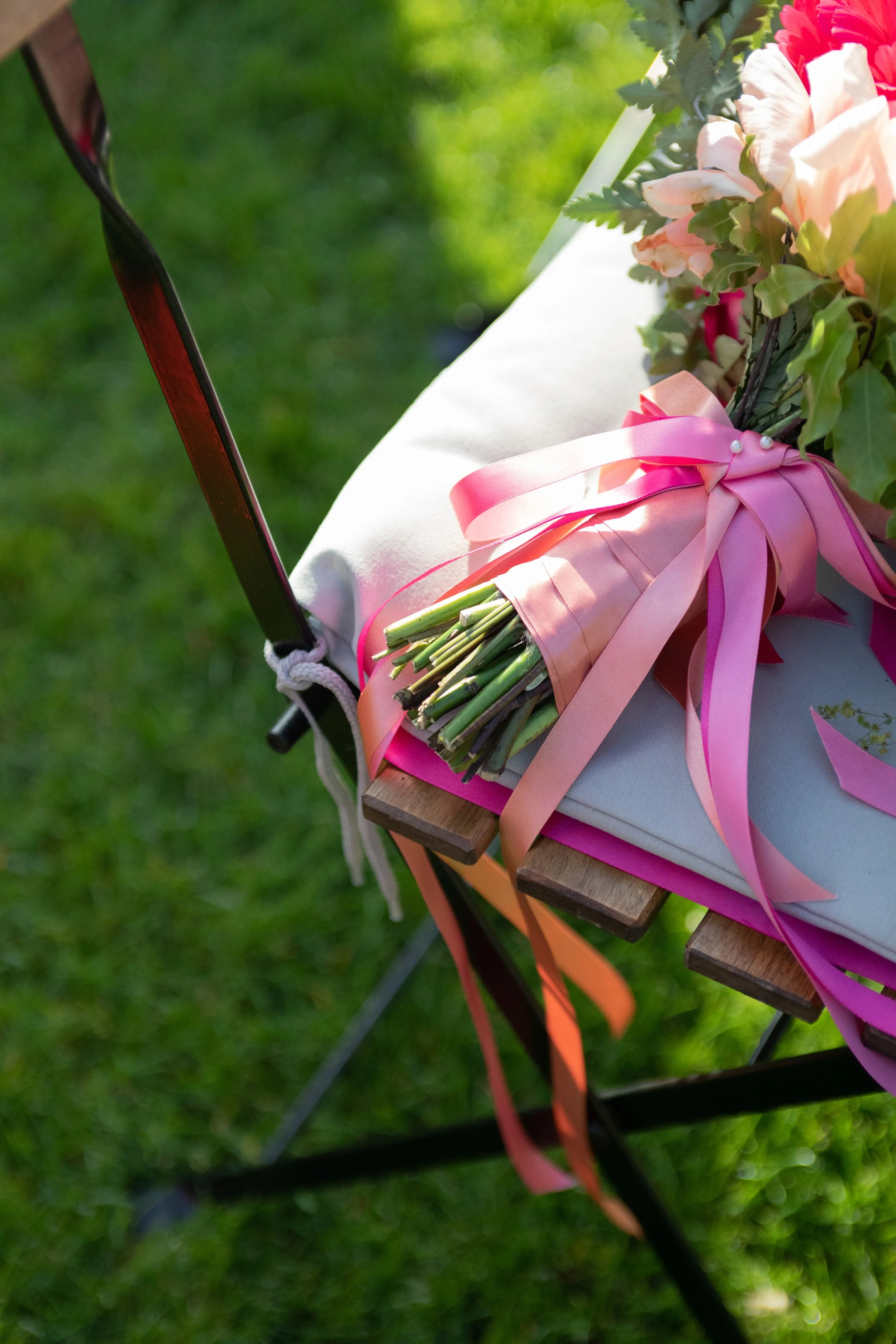 Bouquet of pink and white flowers with green leaves, tied with pink and peach ribbons, placed on a white cloth on a wooden surface, outdoors with green grass background.