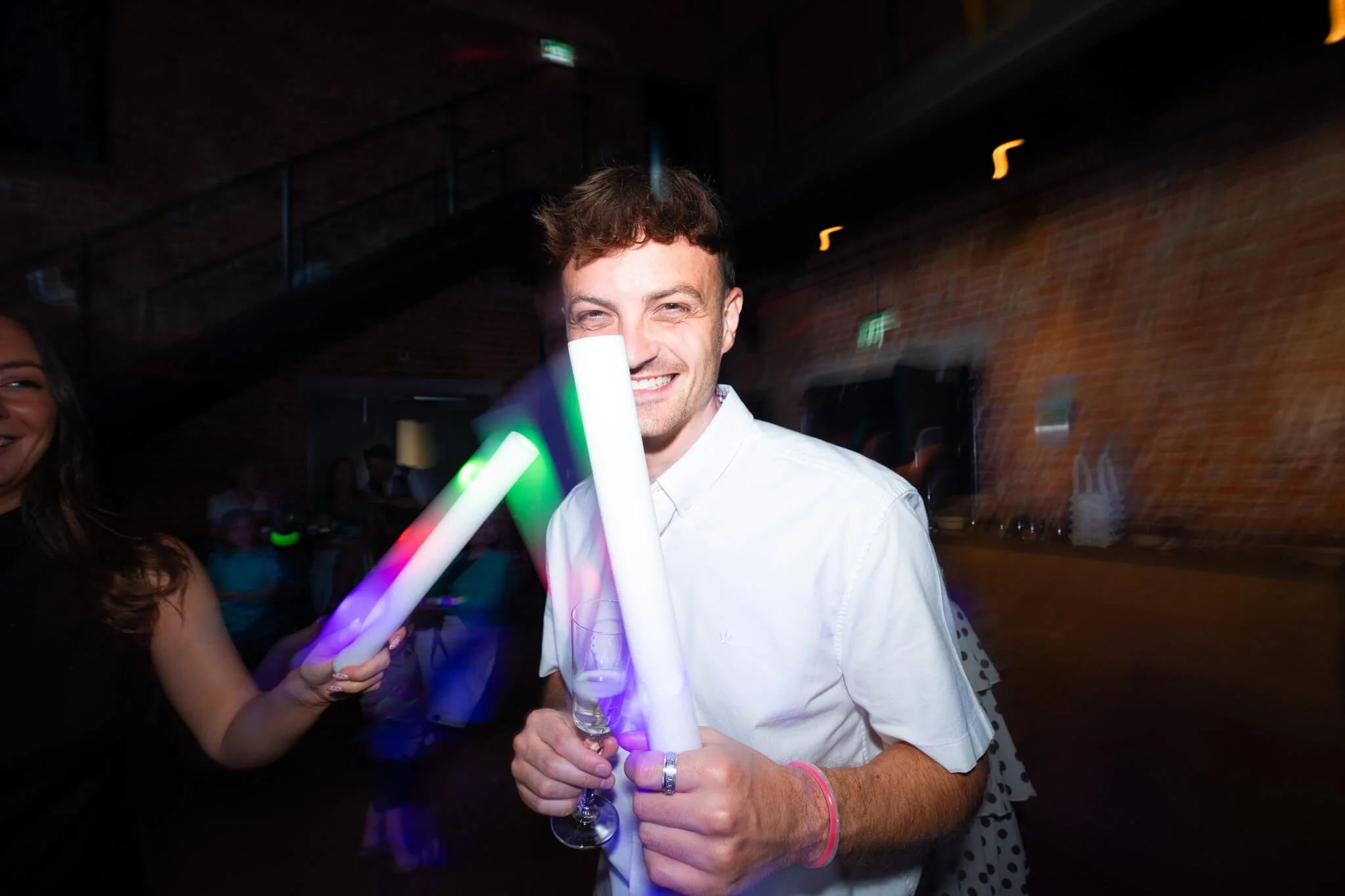 A man smiling at a celebration, holding glow-in-the-dark light sticks and a glass of champagne, in a dimly lit indoor venue with exposed brick walls.