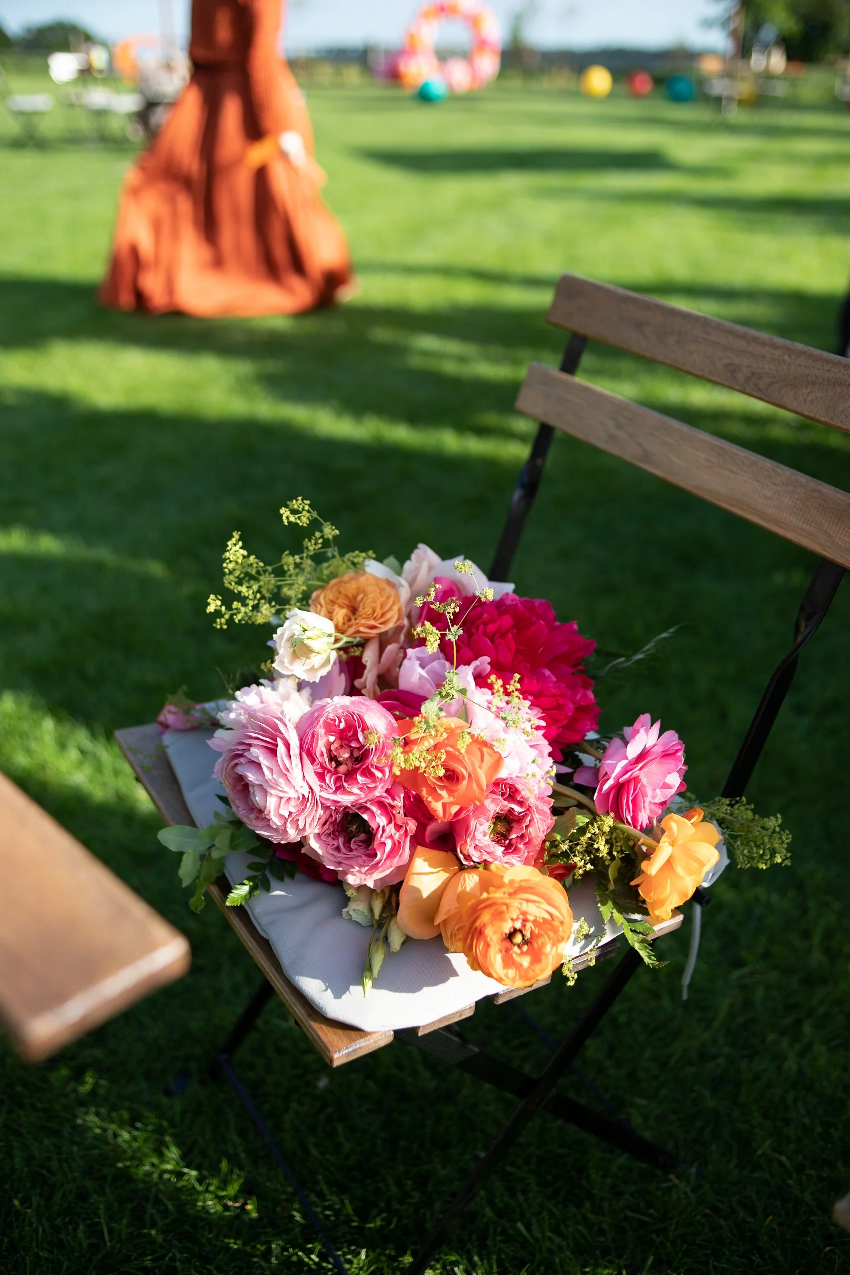 A bouquet of pink, orange, and white flowers on a wooden chair outdoors on a sunny day, with a blurred background of a grassy field and colorful circular objects.
