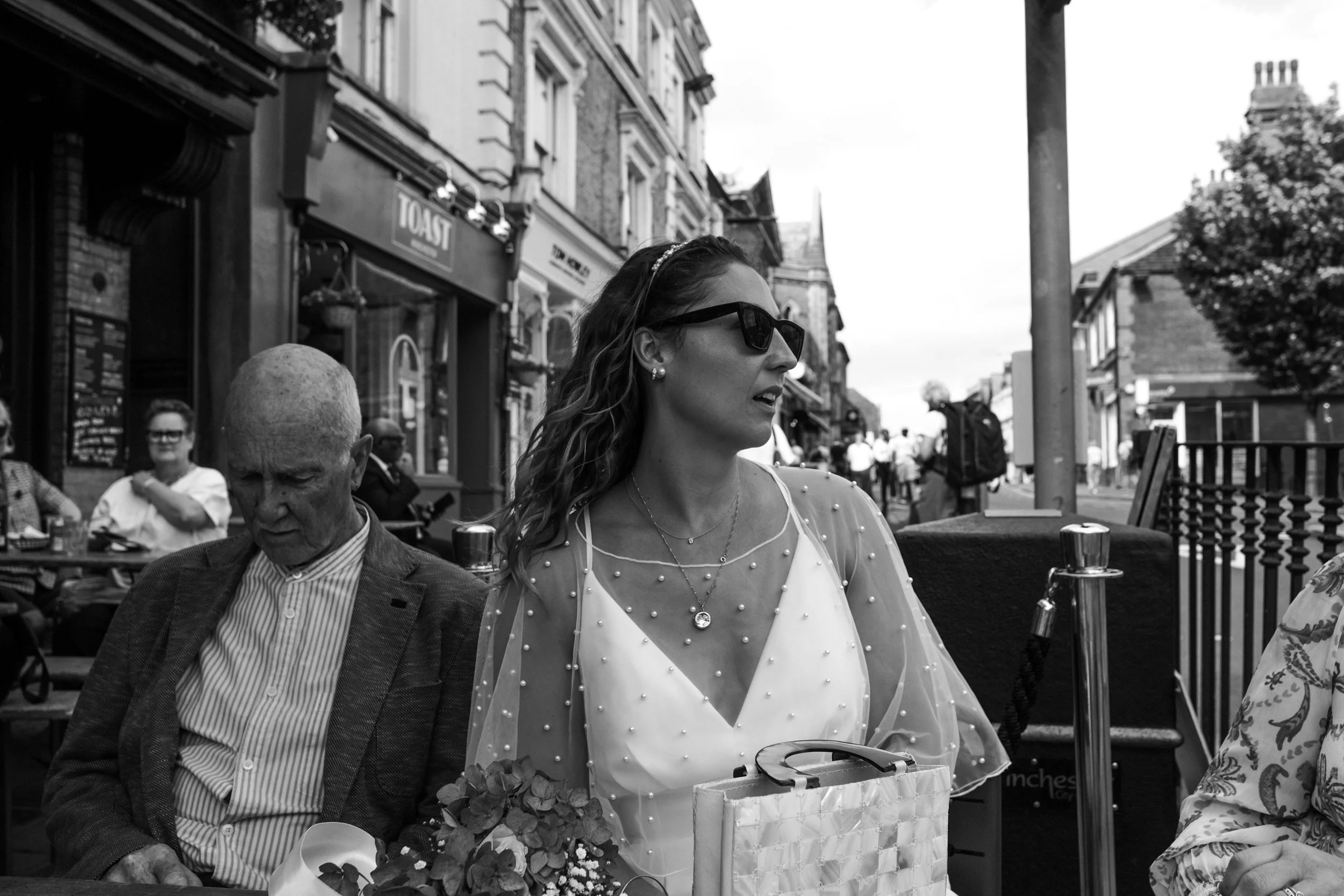 A woman in sunglasses wearing a white dress with pearl embellishments, standing next to an older man in a blazer and striped shirt. They are outdoors in a city street with other people, buildings, and trees in the background.
