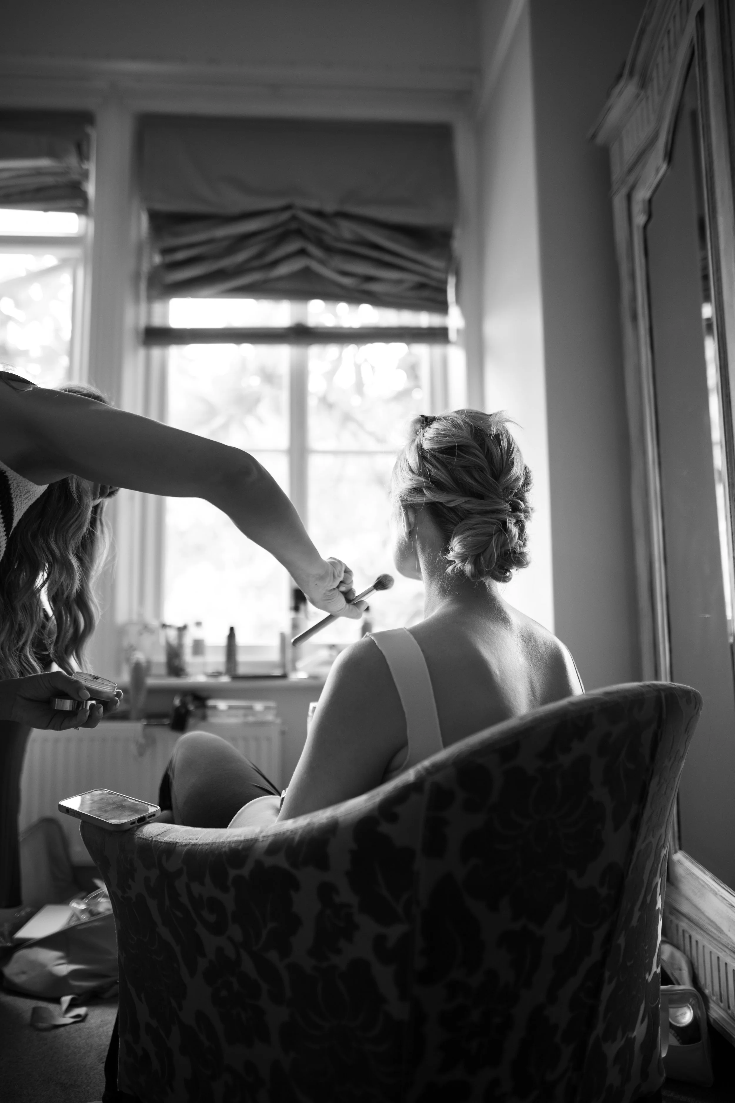 A woman sitting in a vintage armchair with a floral pattern, receiving makeup or hair styling from someone off-camera. The room has large windows with curtains and a dresser with framed pictures or artwork.
