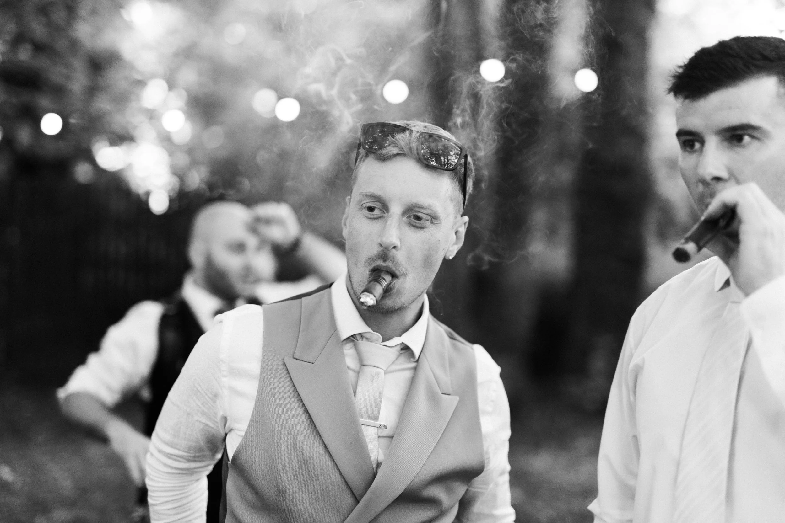 Black and white photo of two men in formal attire, one smoking a cigar. The man in the foreground has sunglasses on his head. People and lights are visible in the blurred background.