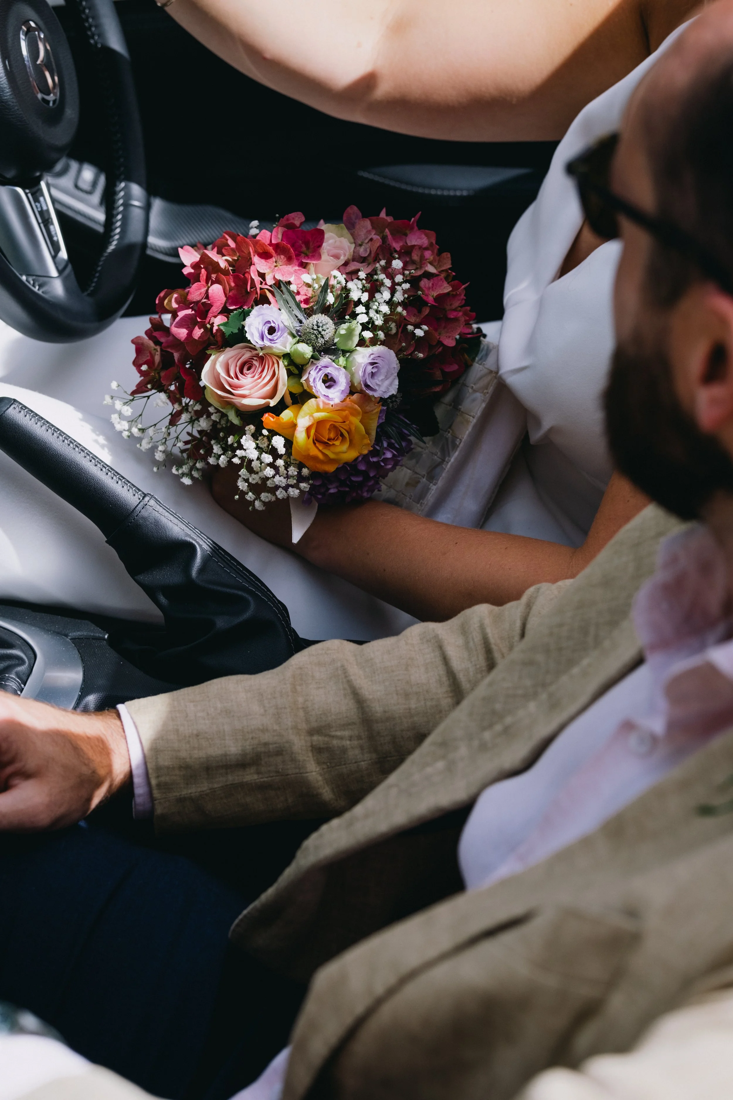 People sitting in a car, with one holding a bouquet of colorful flowers, including roses and baby's breath.