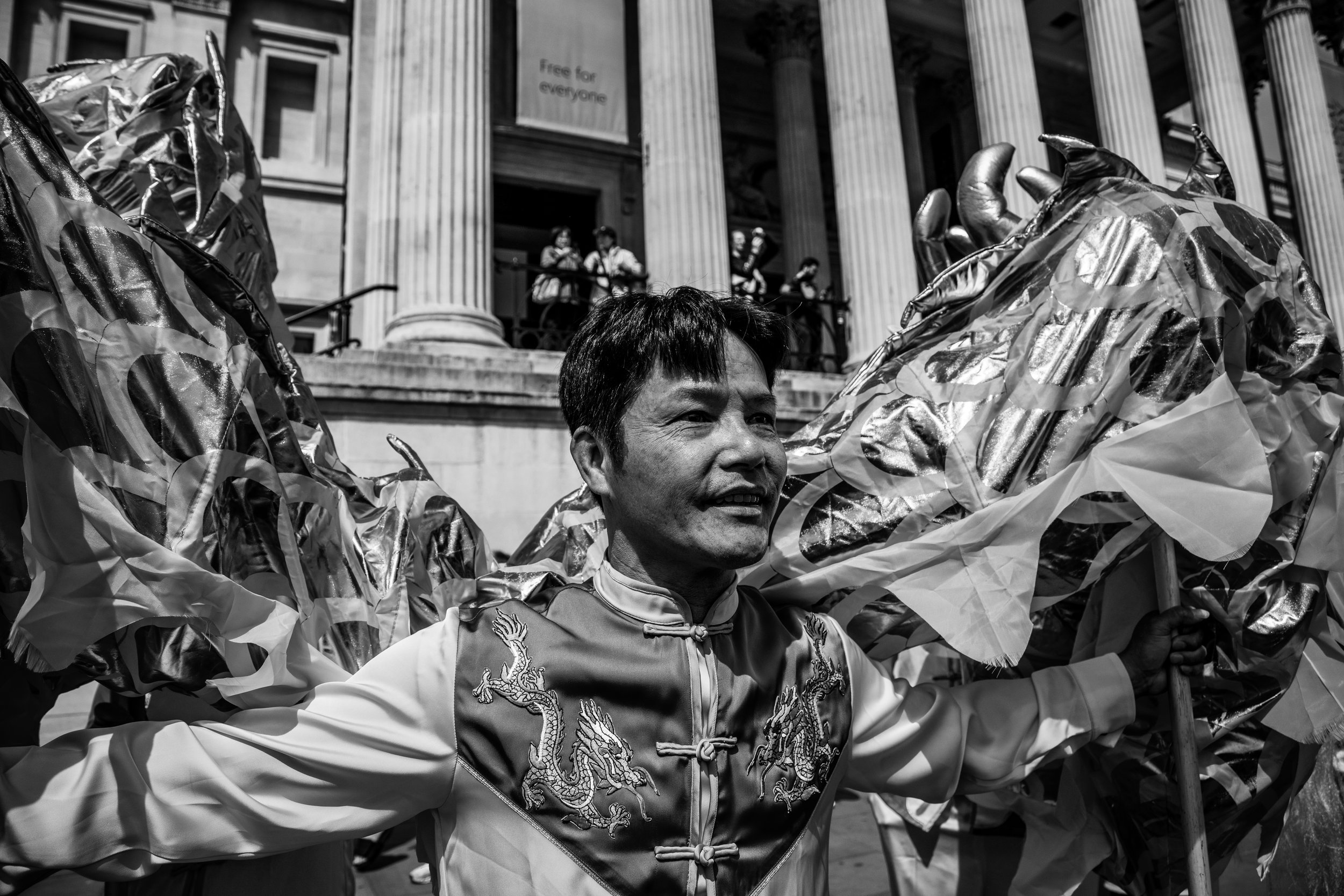A man in traditional Asian attire performing a lion dance with lion costume decorations surrounding him in front of a government building with large columns and a sign that says "free for everyone".